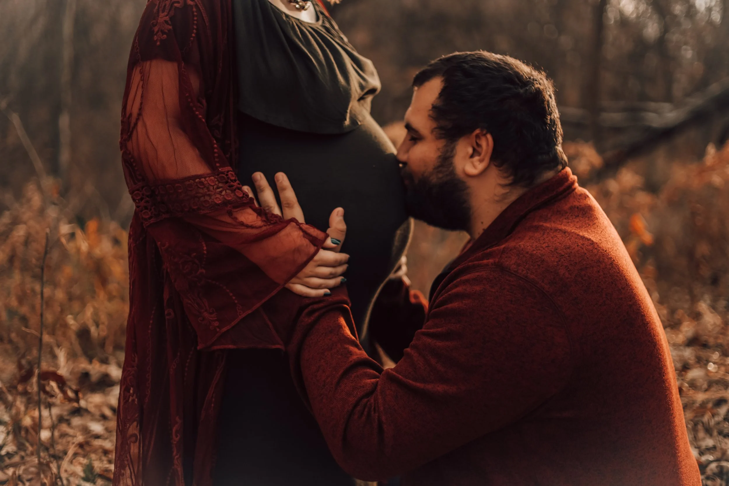 A man kneeling on the ground with his head resting on a pregnant woman's belly, who is standing outdoors in a wooded area during autumn.