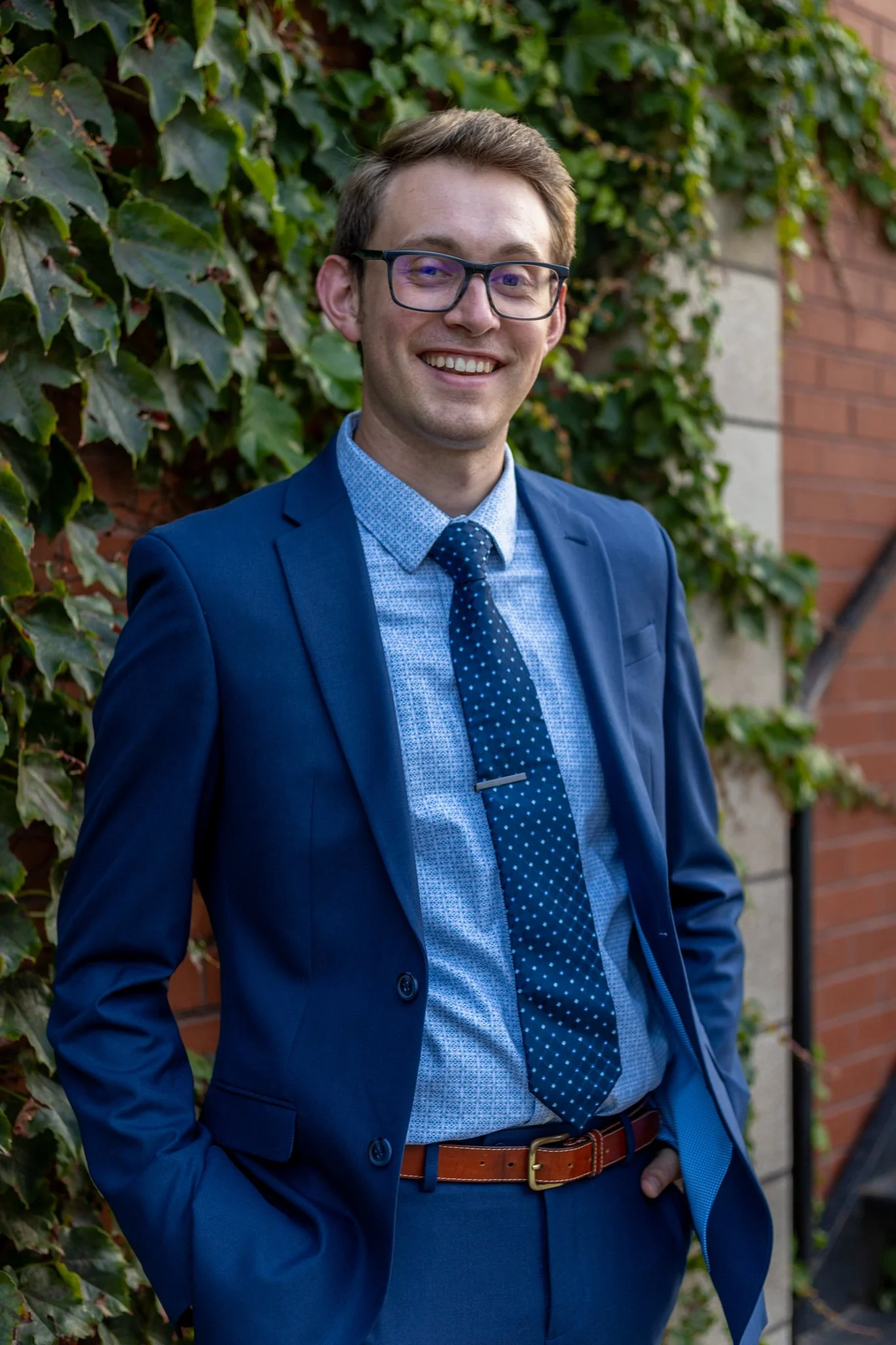 A young man smiling outdoors wearing glasses, a blue suit, a blue patterned shirt, and a blue tie with white dots, standing against a background of green ivy and a brick wall.