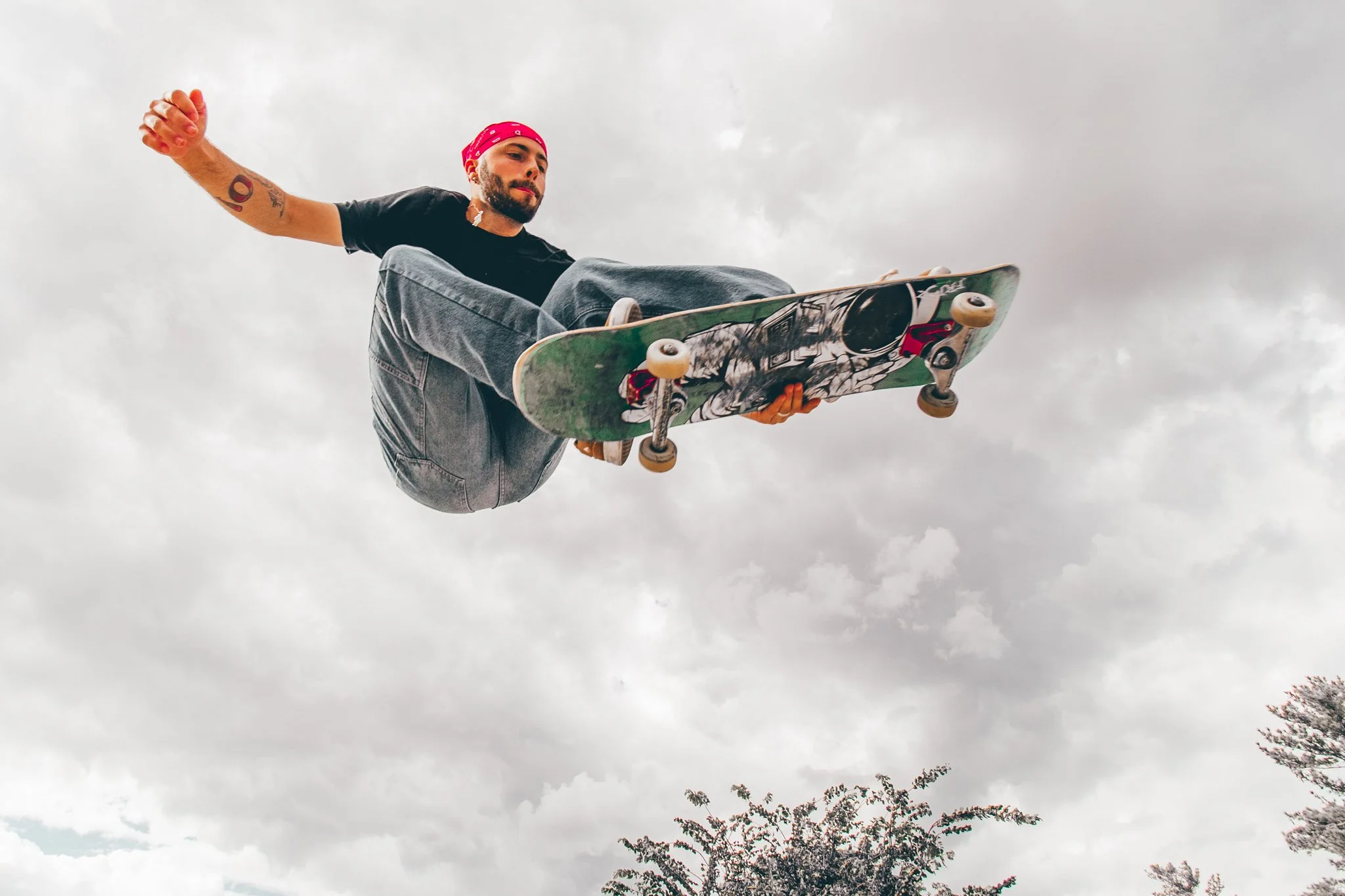 A person skateboarding in mid-air against a cloudy sky, wearing a pink bandana, black t-shirt, and gray pants.
