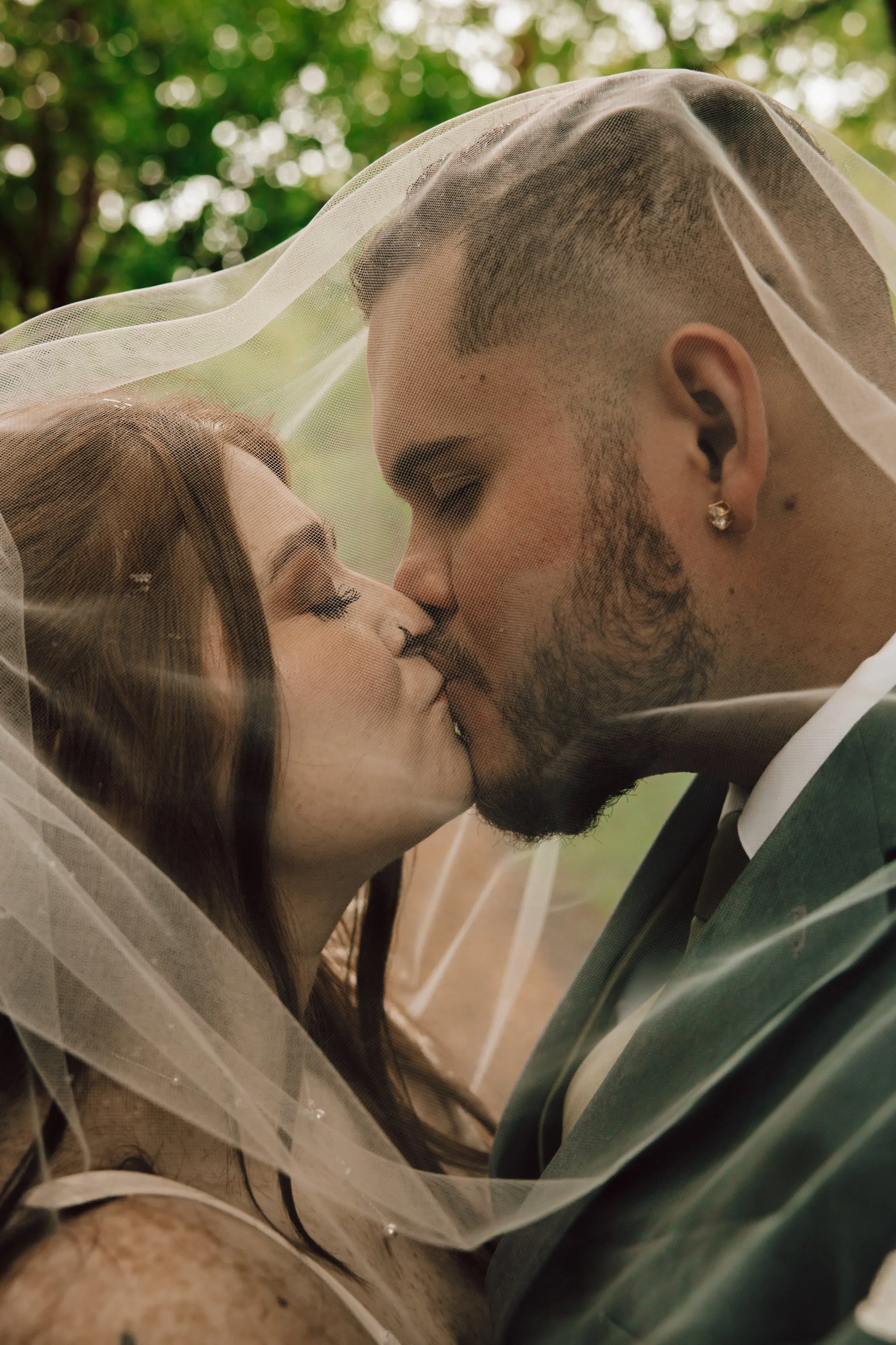 A couple leaning close, protected by a sheer veil, outdoors with green trees in the background.
