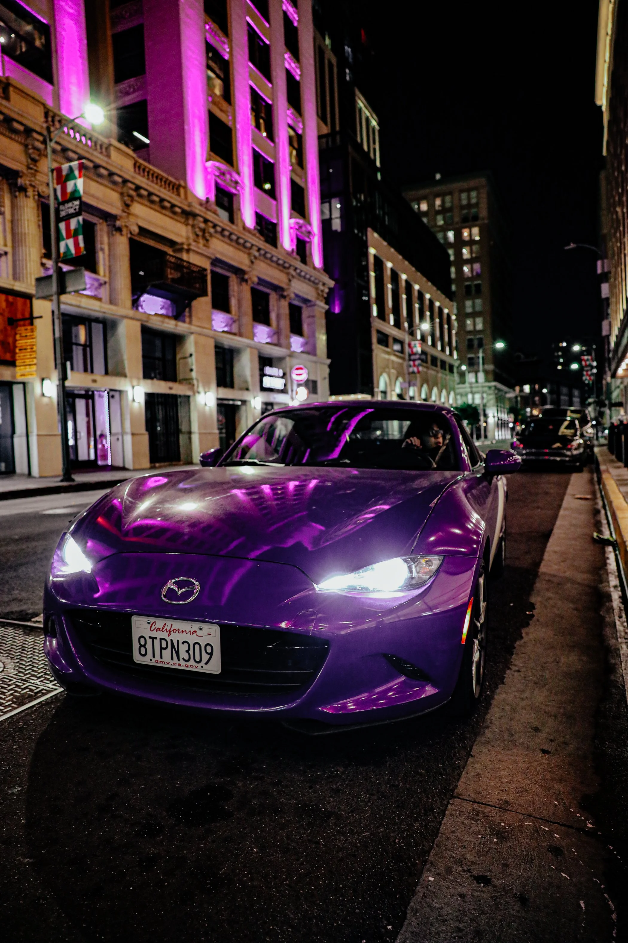 A purple Mazda sports car with California license plate 8TPN309 parked on city street at night, reflected purple lights on the car, tall buildings with purple lighting, and streetlights in the background.