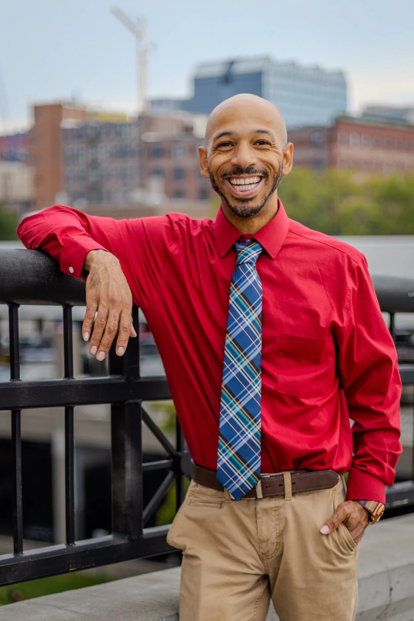 A man with a shaved head, beard, and mustache smiling, wearing a red shirt, plaid tie, and khaki pants, leaning on a black railing with an urban background of buildings and a crane.