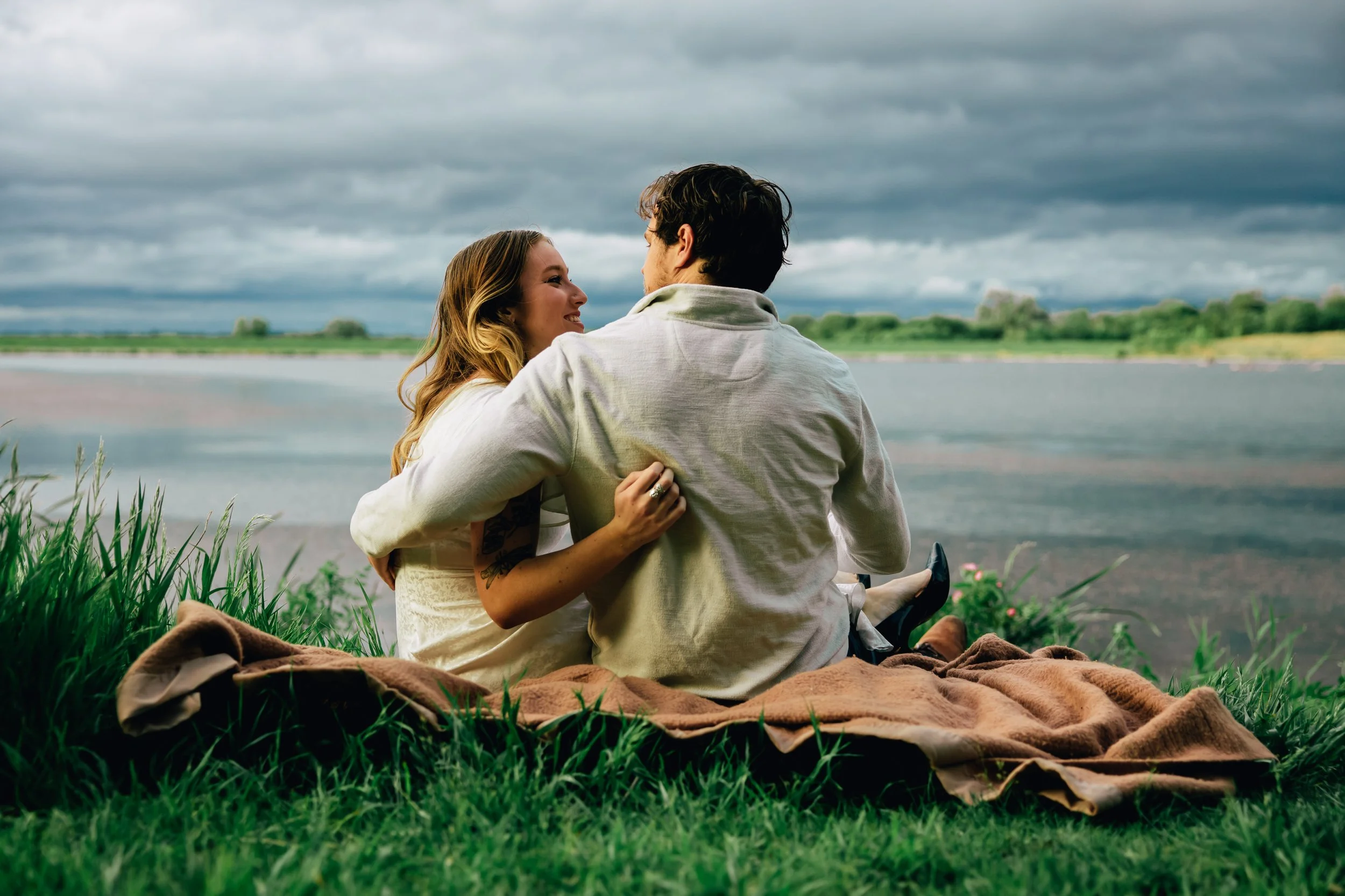 A couple sitting on a blanket by a lake during cloudy weather, facing each other and smiling.