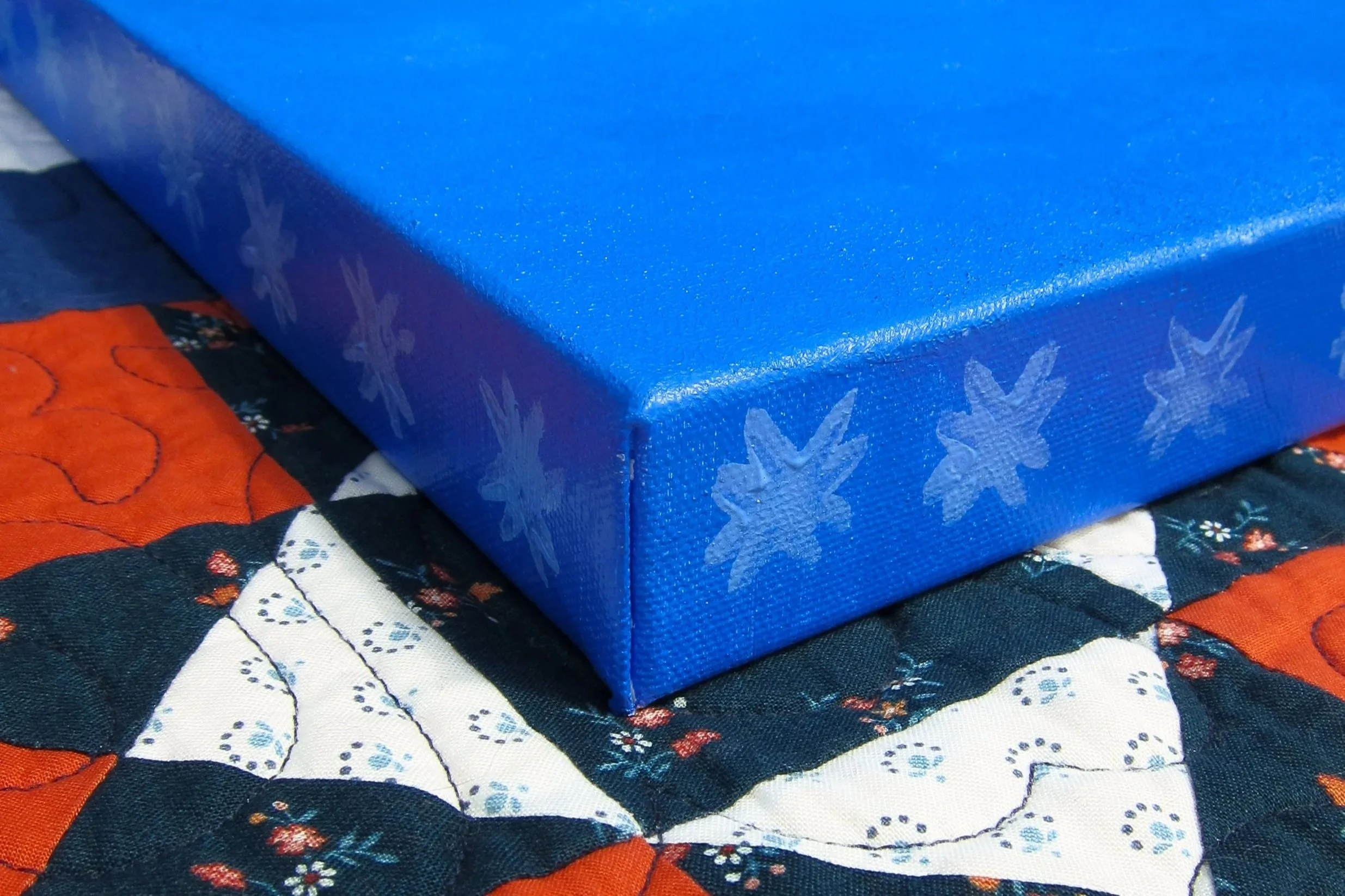 Close-up of a blue gift box with a snowflake pattern on the side, placed on a quilted fabric with a floral and geometric pattern.