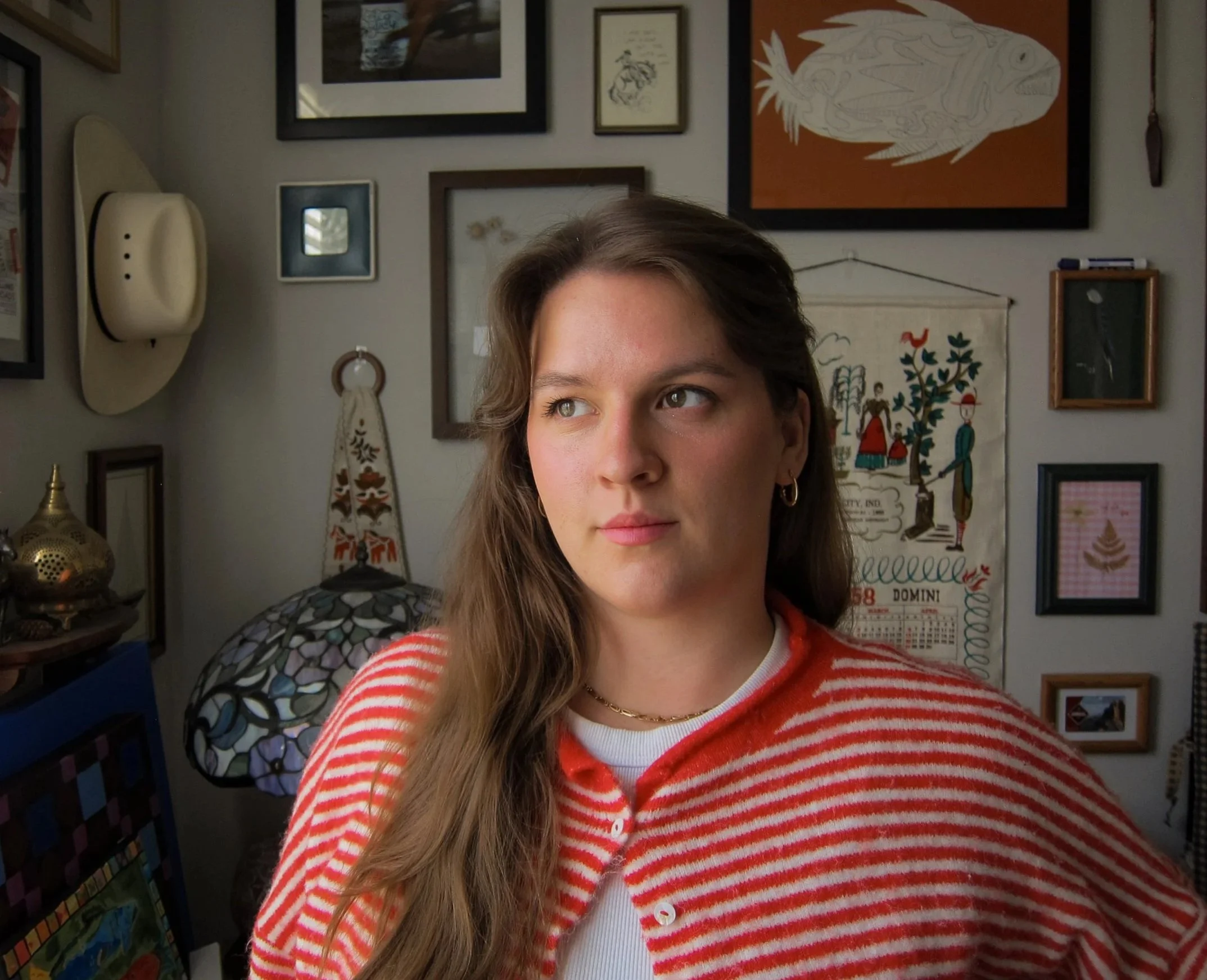 Close-up of a woman with long brown hair, wearing a red and white striped sweater and gold hoop earrings, standing in front of a wall decorated with framed pictures and artwork.