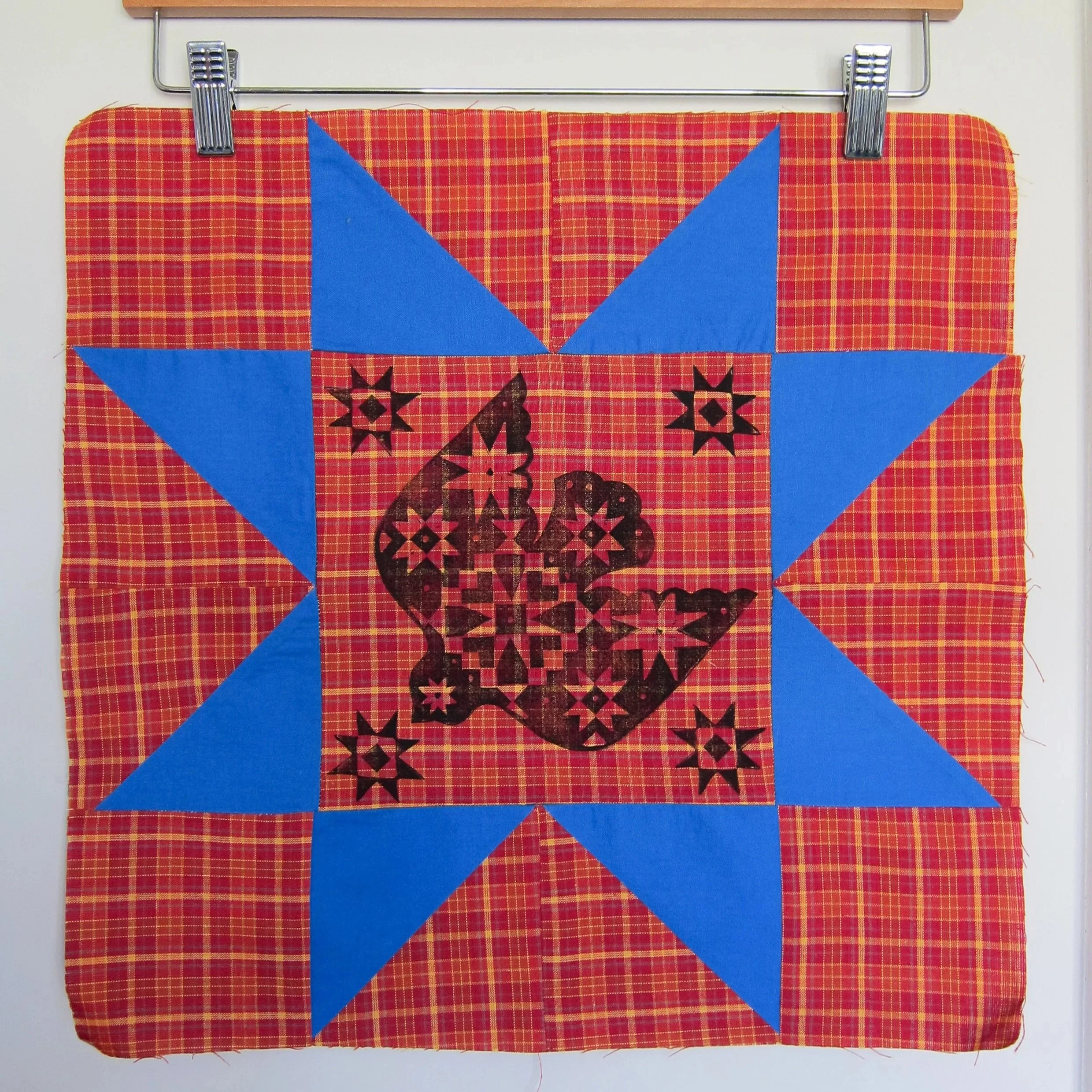 Close-up of a quilt with a red and yellow plaid background, blue fabric triangles, black star-shaped patterns, and the text "Merry Christmas" in black letters.