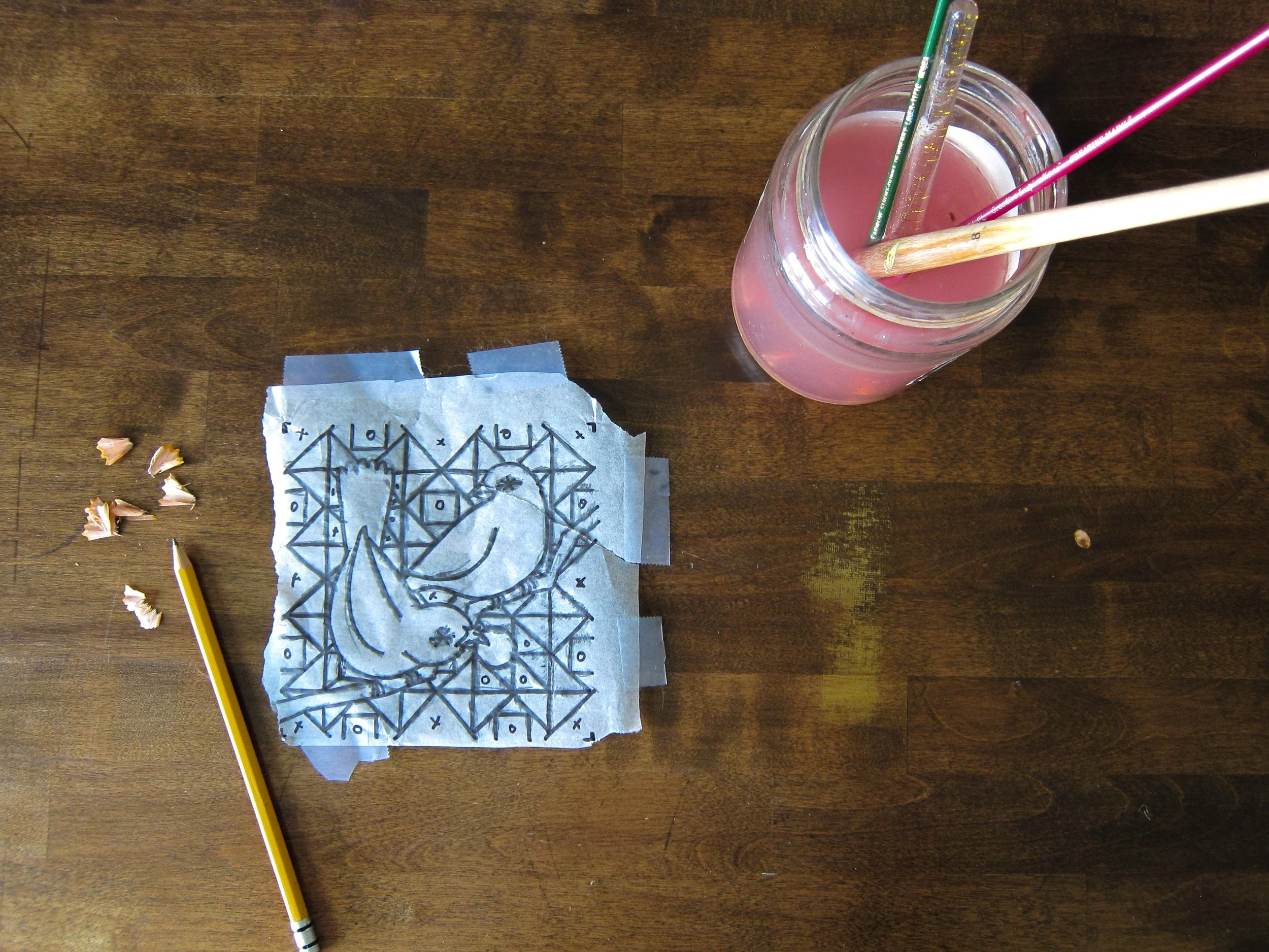 A glass jar filled with pink liquid containing three straws on a wooden table. There are pencil shavings, a yellow pencil, and a piece of paper with a geometric drawing.