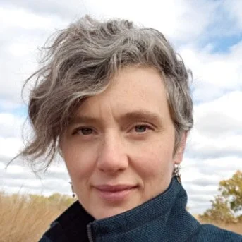 Portrait of Alina Campana outdoors with cropped flowing gray hair in a side part and wearing a pulled up blue collar
