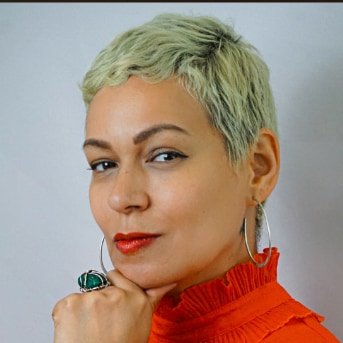 Woman with short cropped blonde hair wearing a bright orange high-neck shirt, big hoop earrings, and resting chin on hand.