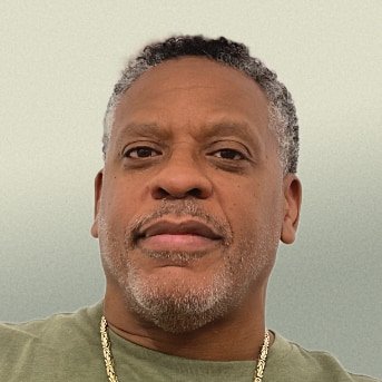 Portrait of a black man with salt and pepper goatee wearing a olive green shirt and a gold necklace 