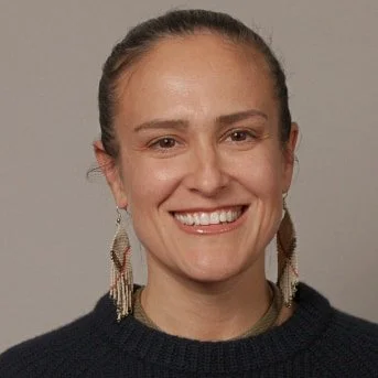 Close-up of a woman smiling, wearing earrings and a dark sweater against a neutral background.
