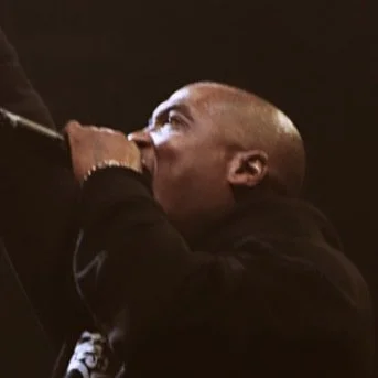 A man with a shaved head singing into a microphone on stage.