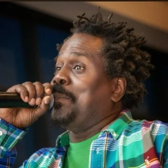 Man with curly hair singing into a microphone, wearing a colorful plaid shirt and a green t-shirt.