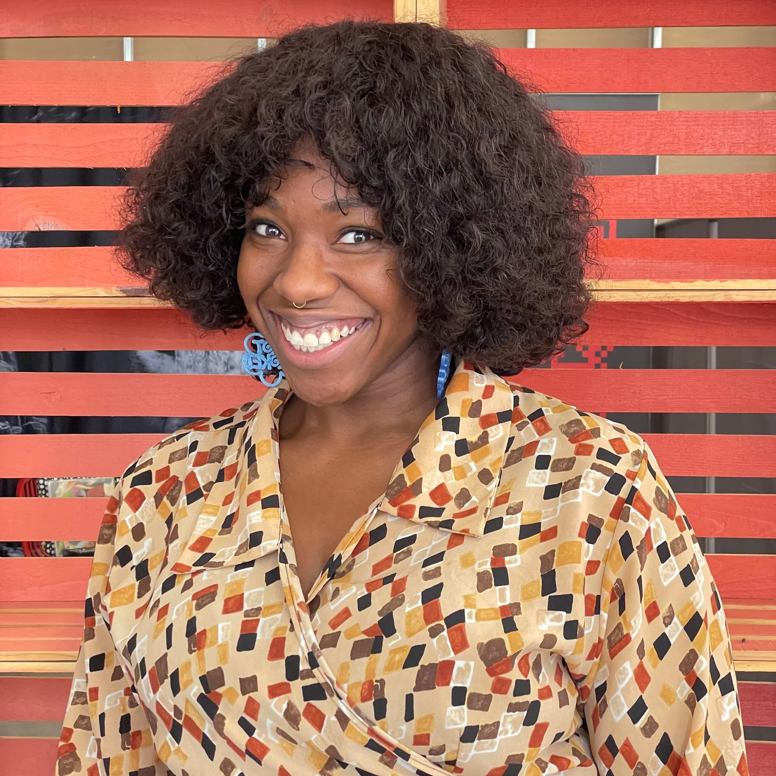 A woman with curly hair and blue earrings smiling in front of a red wooden slat background, wearing a patterned shirt.
