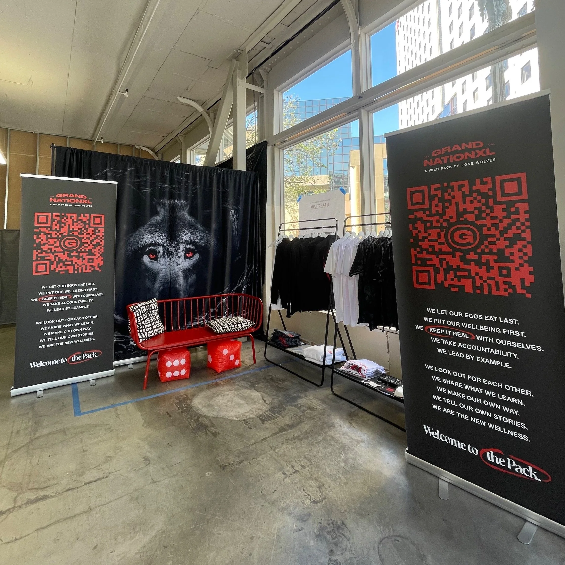 Display booth with banners featuring a lion's face and text about a pack, with clothing on racks, a red bench, and dice-shaped stools in an indoor setting.