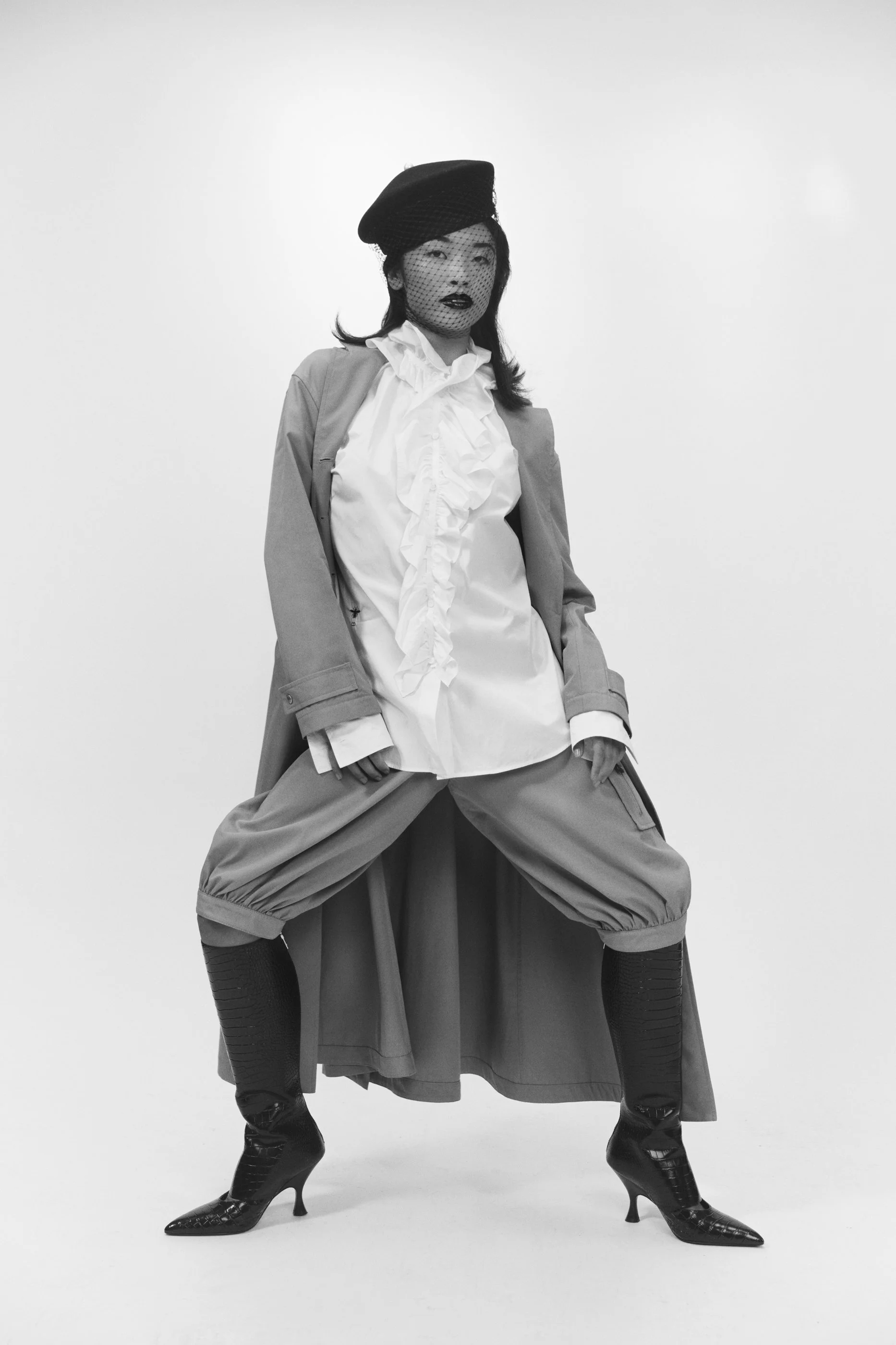 American Ballet Theater dancer Kayla Mak with a full Christian Dior look including a hat with vail over her face, a ruffled blouse, a blazer, wide-legged riding pants, and high-heeled boots, posing with legs apart against a white background.