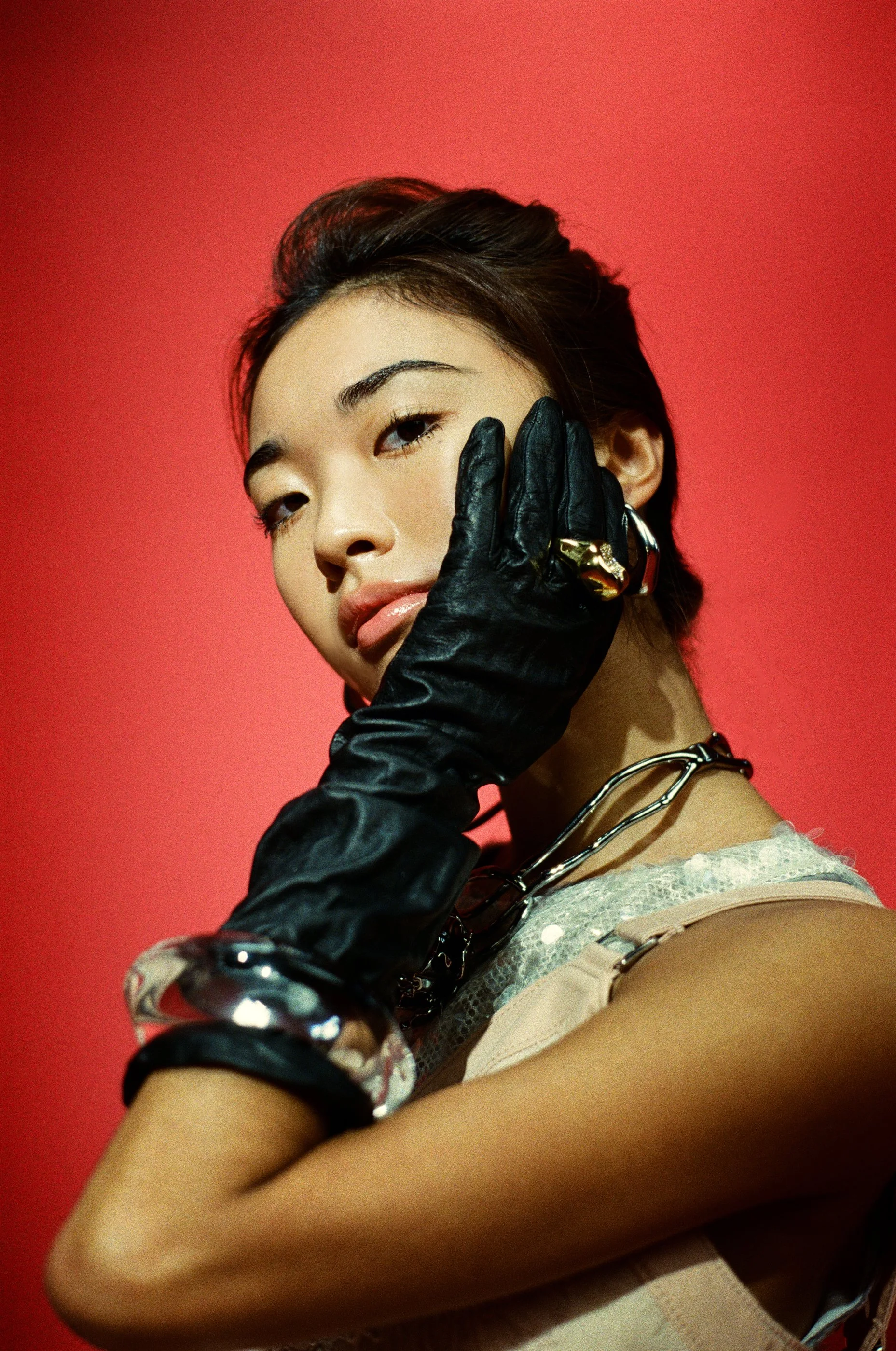 Kayla Mak, a ballet dancer for ABT in New York City, wearing a silver sequin 3.1 Phillip Lim dress, poses against a red background. She is accessorized with rings and necklaces by Alexis Bittar worn over black leather gloves for The Glass Magazine.
