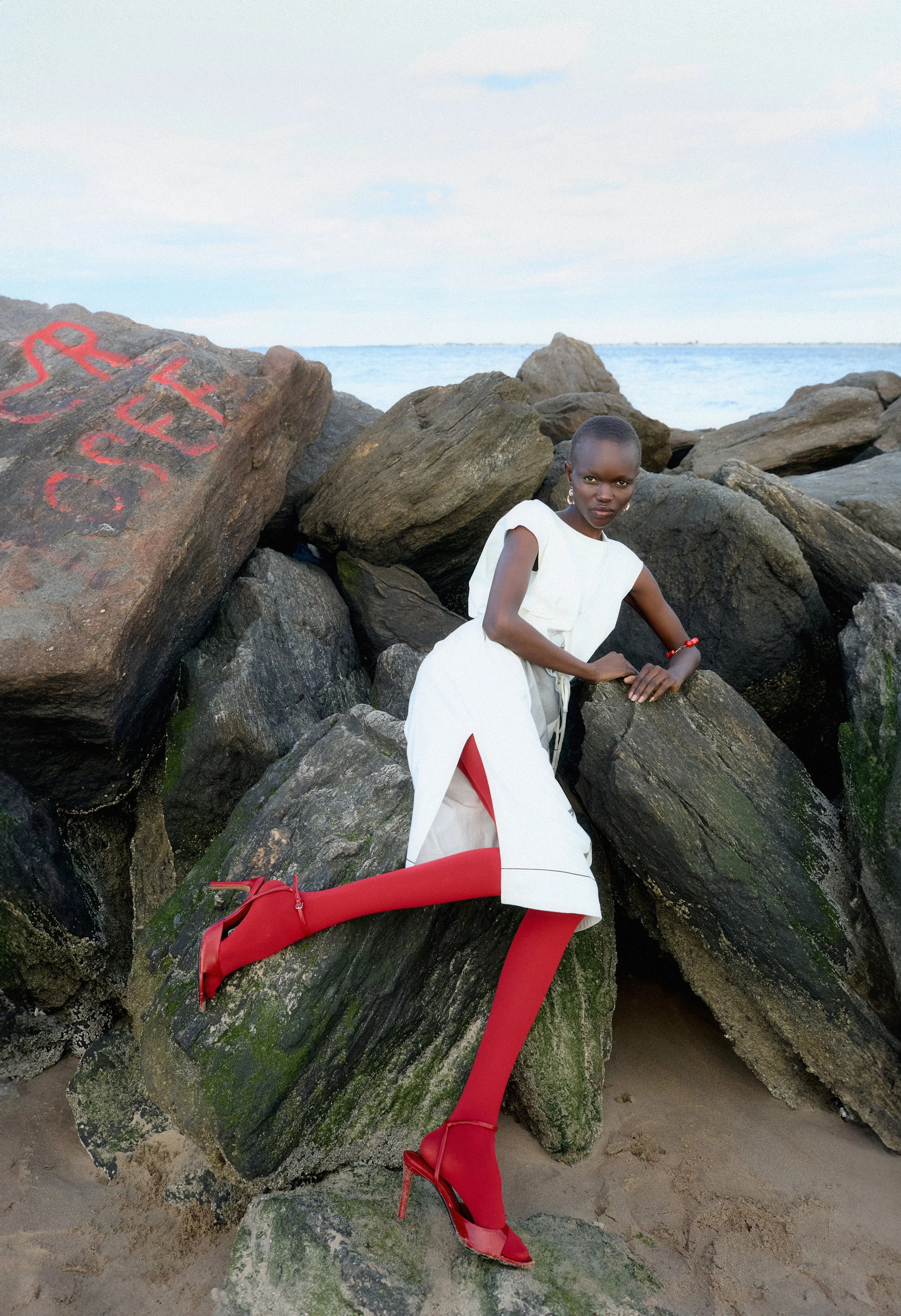 Still photograph of model Adau Aguer where she is laying on rocks in a white dress at Brighton Beach Brooklyn directed by Fashion Filmmakers Indie Studios and photographed by Alessandro Lo Faro
