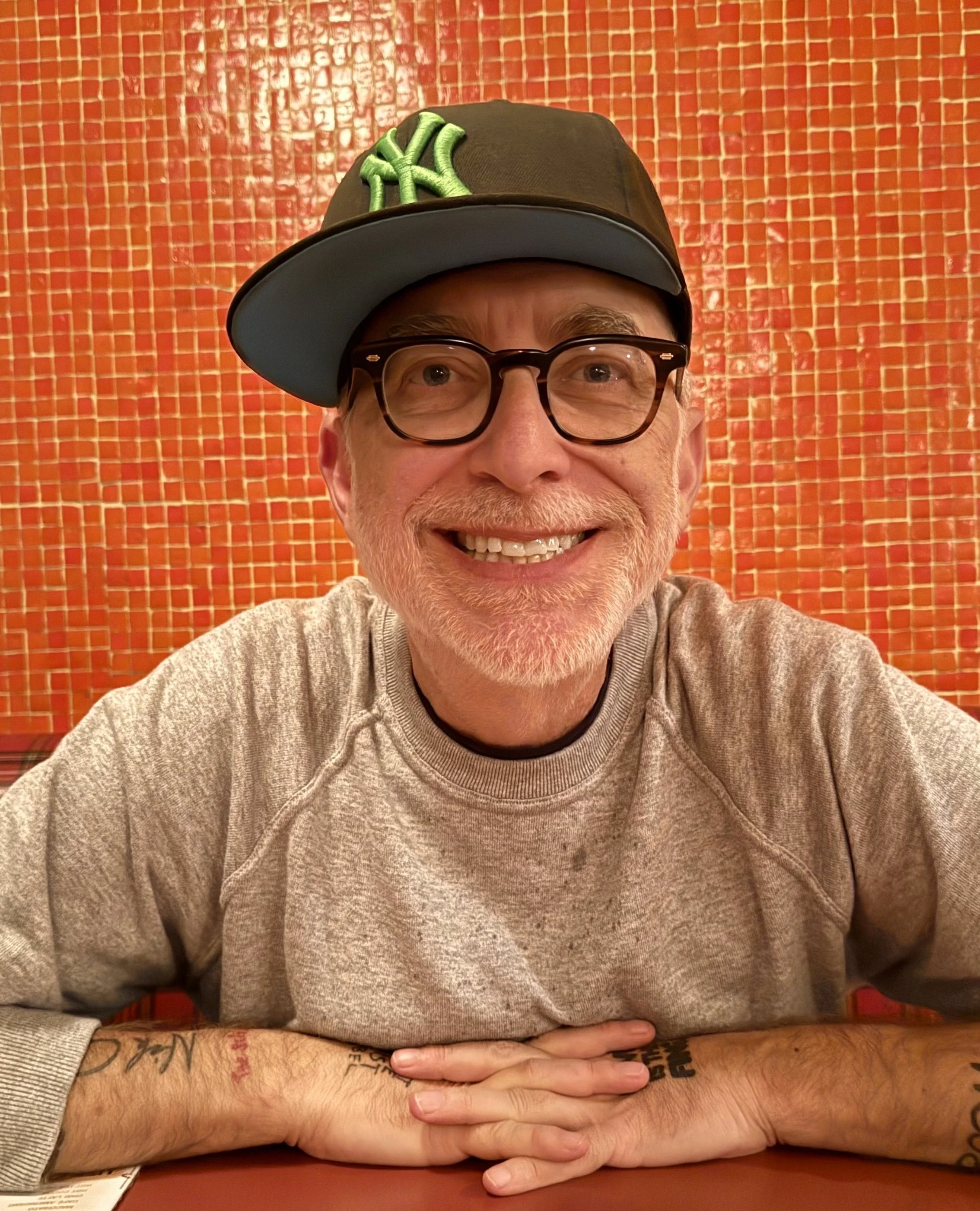 Aaron Blitzstein, seated at a restaurant table, wearing glasses and a baseball cap with a green NY logo, against an orange tiled wall.