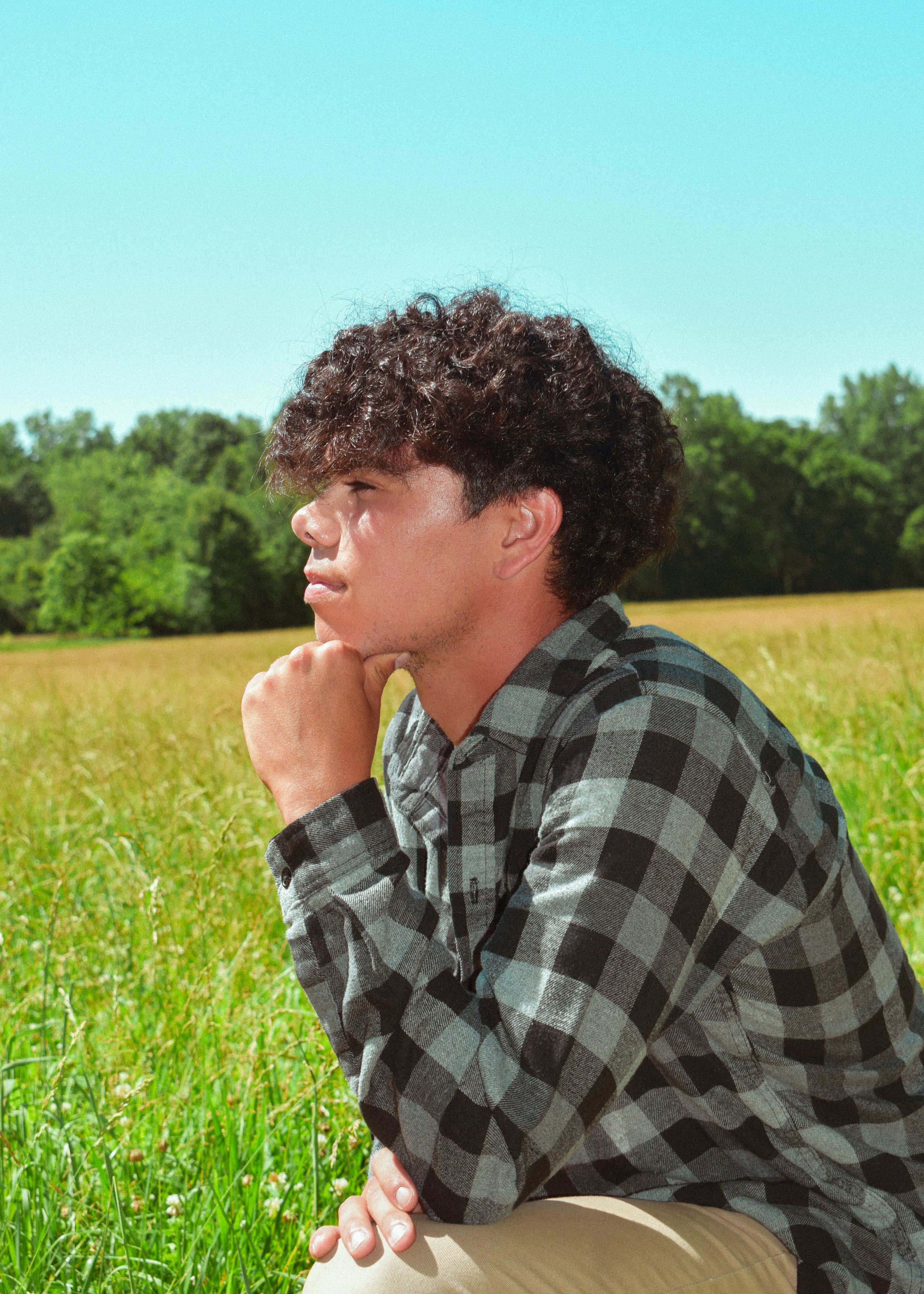 A young man with dark curly hair sitting in a grassy field under a clear blue sky, wearing a black and gray plaid shirt, resting his chin on his fist and looking to the side.