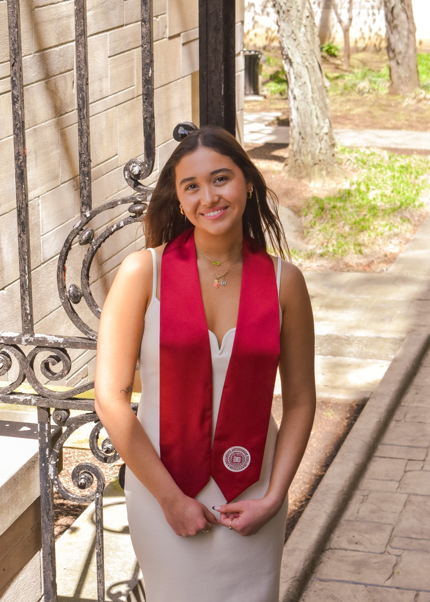 Young woman in a white dress with a red graduation stole, standing outdoors near a brick wall and decorative iron fence, smiling, with trees and sidewalk in the background.