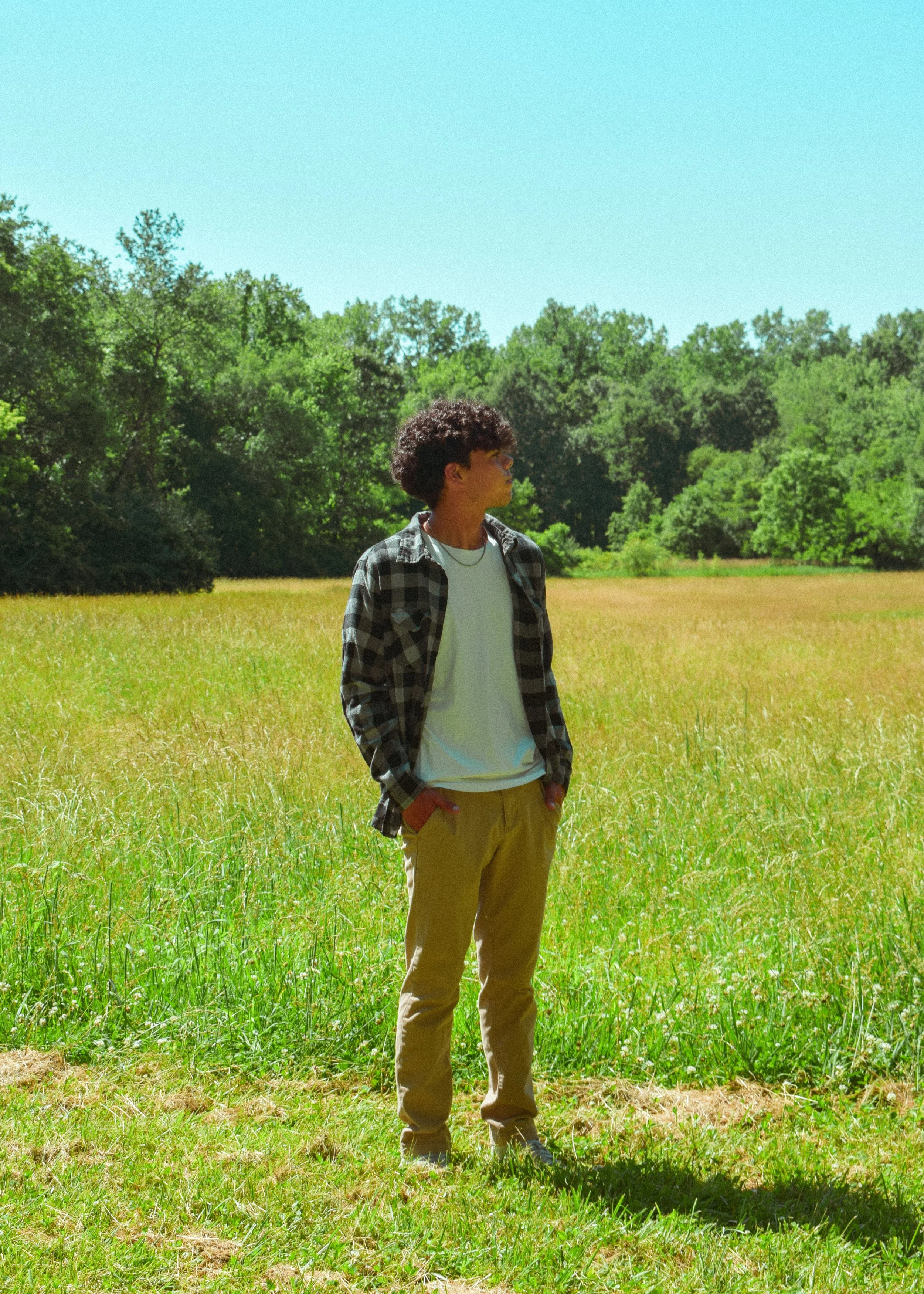 A young man standing in a grassy field with his hands in his pockets, wearing a black and gray checkered shirt, white t-shirt, and khaki pants. He has curly hair and sunglasses, gazing to the side, with trees and blue sky in the background.