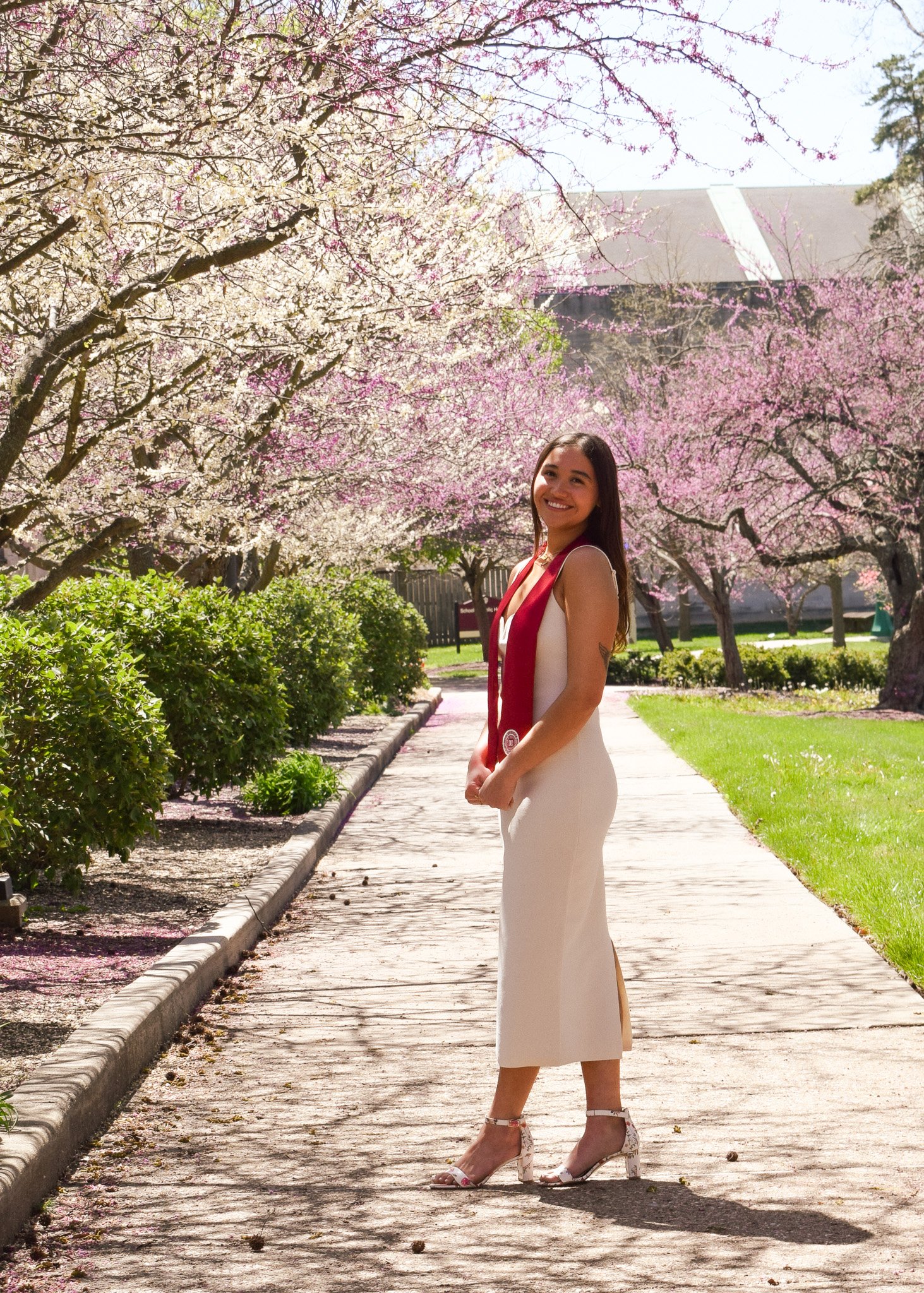 A woman in a white dress and heels standing on a paved walkway lined with bushes, smiling, with pink and white blossoming trees and a building in the background.
