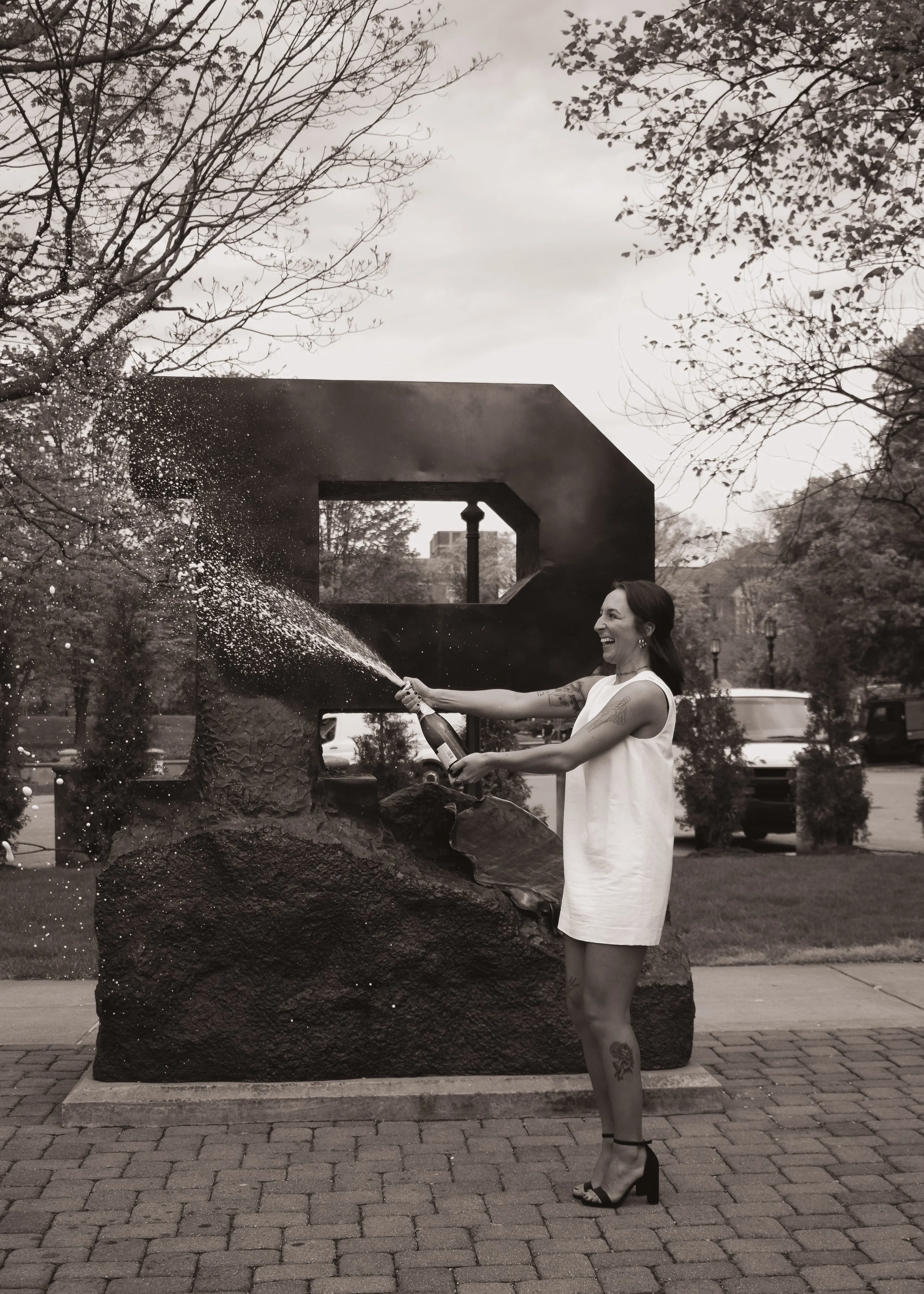 A woman in a sleeveless white dress and high heels is holding a champagne bottle, spraying it in celebration, outdoors in front of a large sculpture and trees.