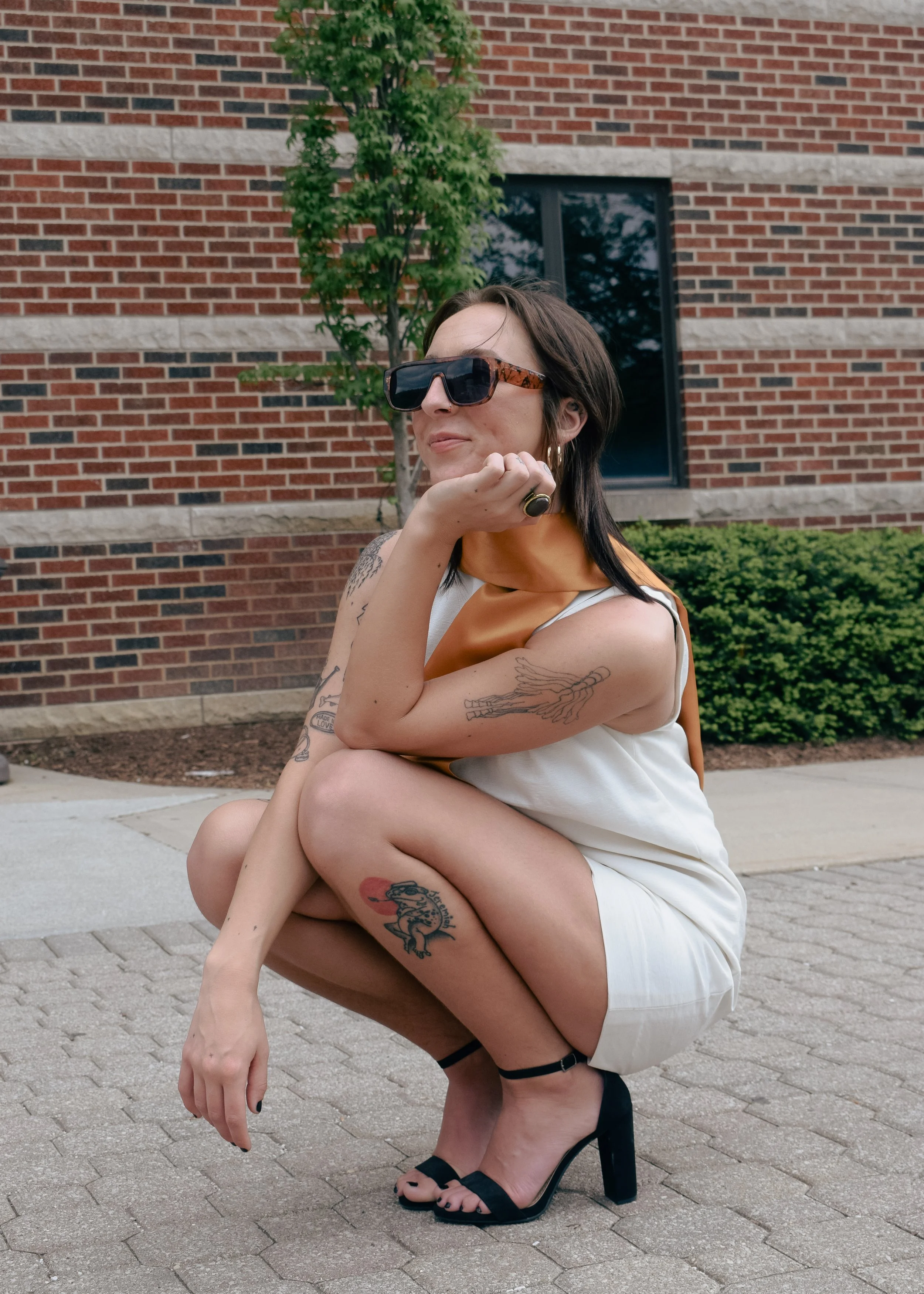 A woman with dark hair, wearing sunglasses, a sleeveless dress, and black high-heeled sandals, squatting on a sidewalk in front of a brick building with greenery.