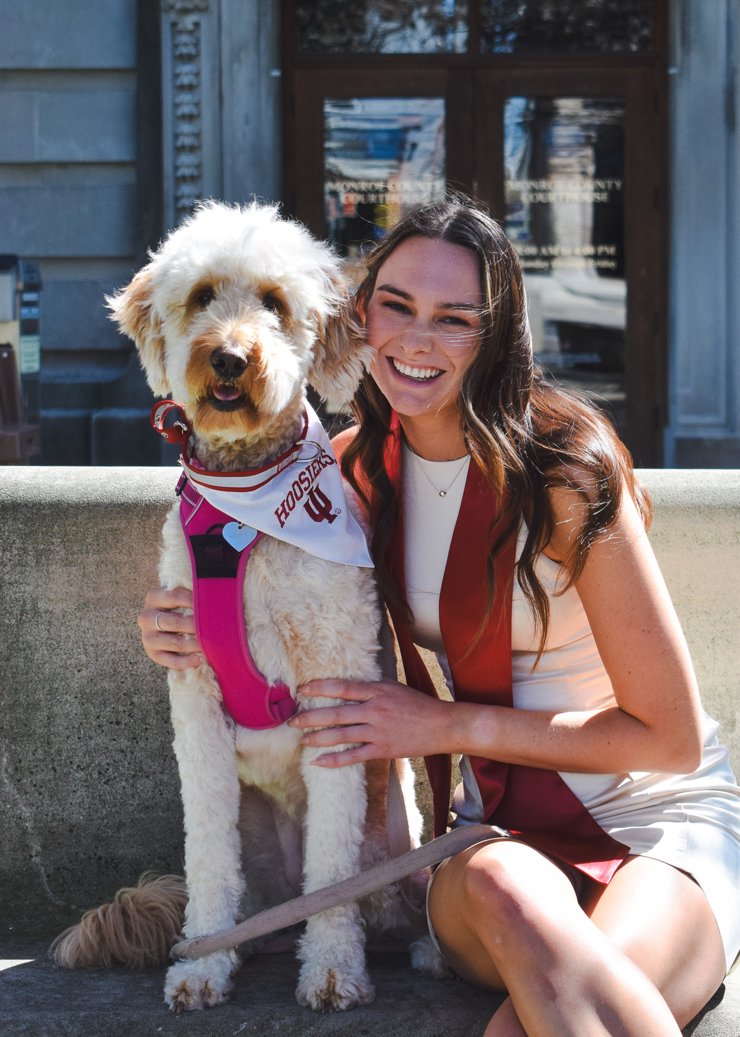 A woman smiling while sitting next to a Labrador retriever wearing a red and white North Carolina bandana and a pink harness, outdoors on a sunny day.
