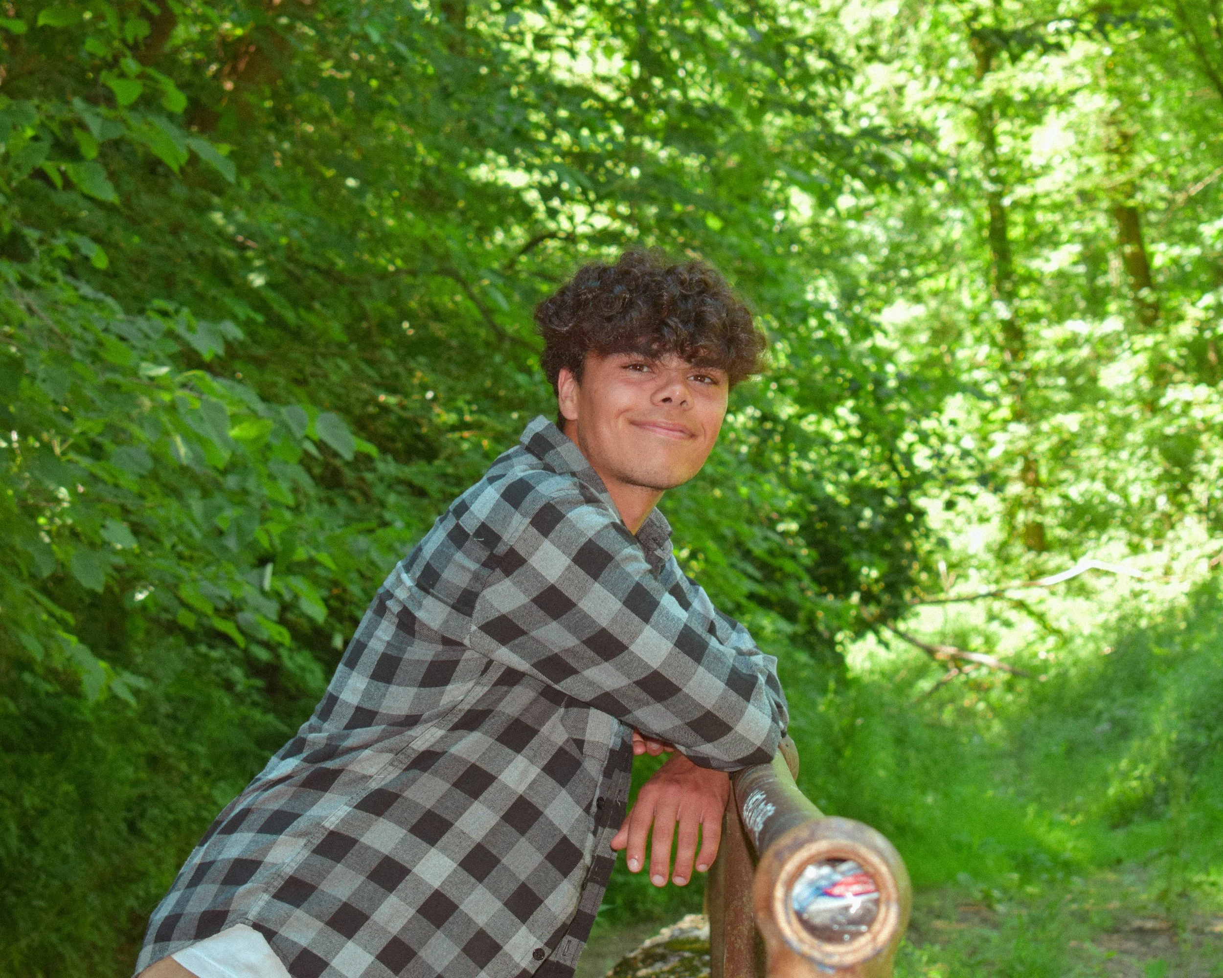 A young man with curly hair standing outdoors in a lush green forest, leaning on a wooden railing and smiling at the camera.