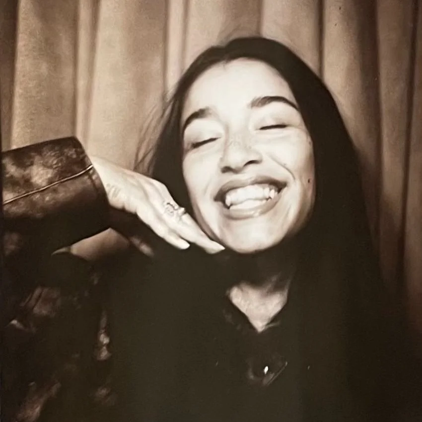 A woman smiling,with her hand resting under her face, in front of a fabric background.