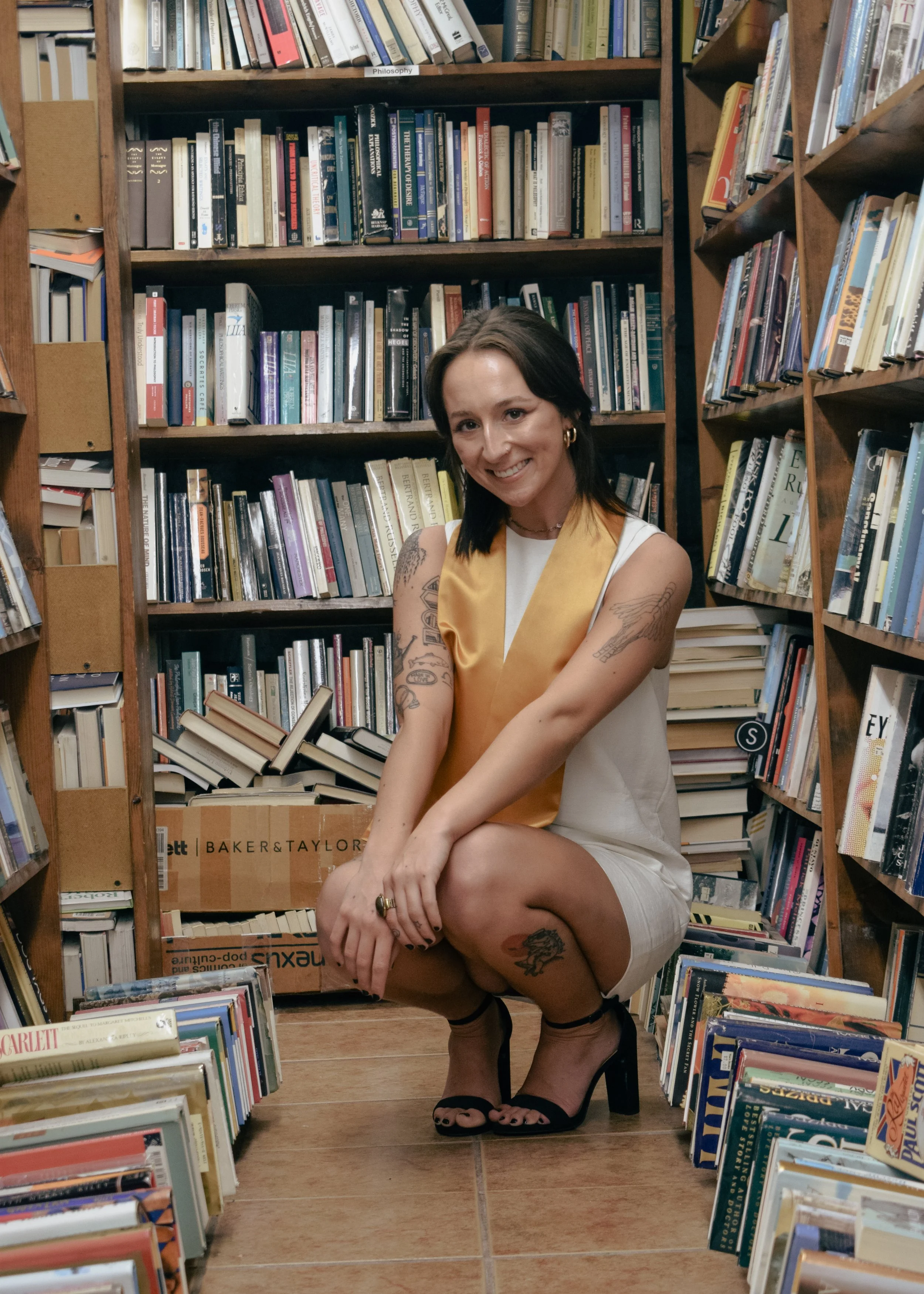A woman squatting in a bookstore aisle, smiling, surrounded by shelves filled with books.