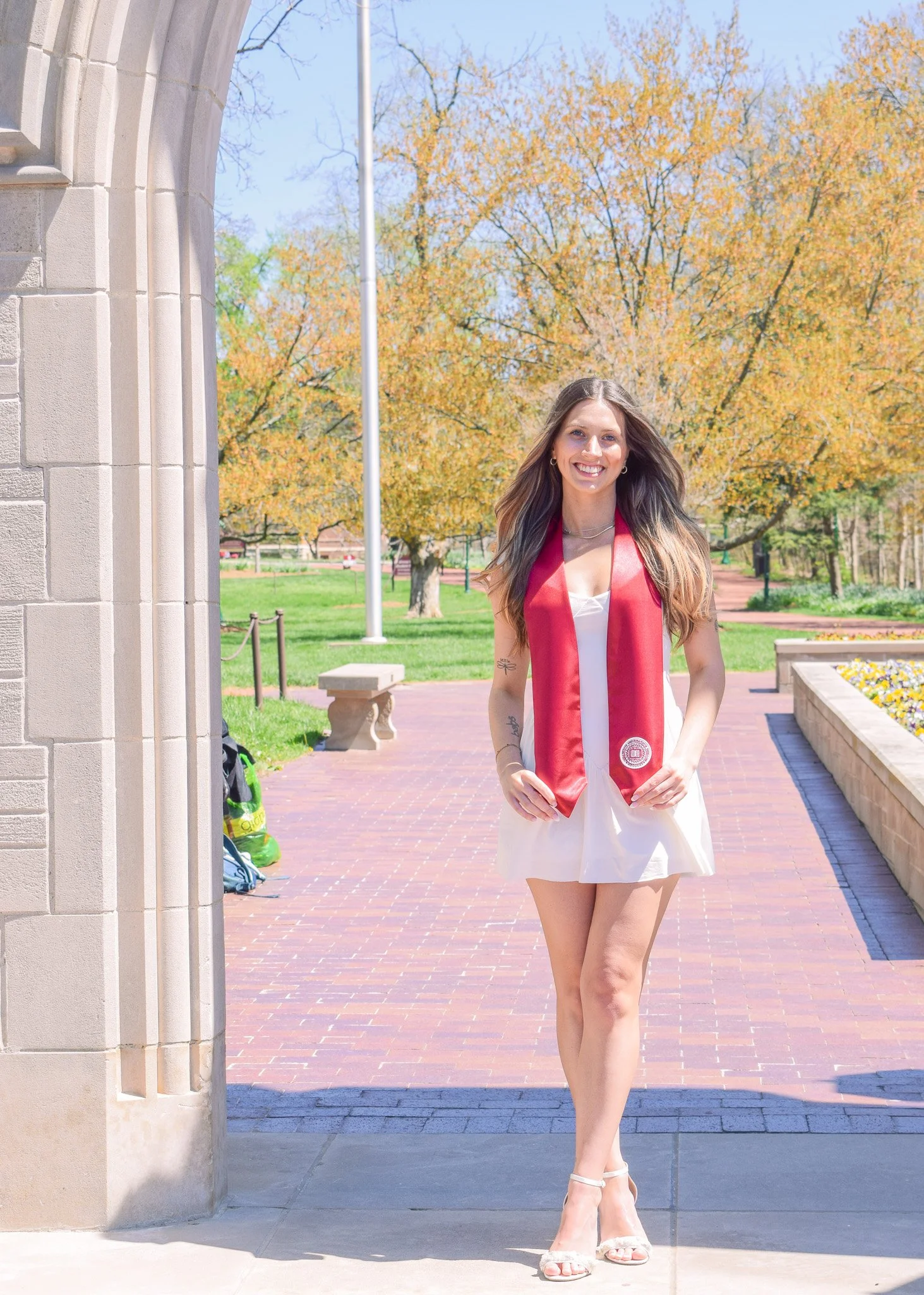 A young woman in a white dress and high heels stands in a park during graduation day, wearing a red graduation stole, smiling in front of trees with autumn leaves.