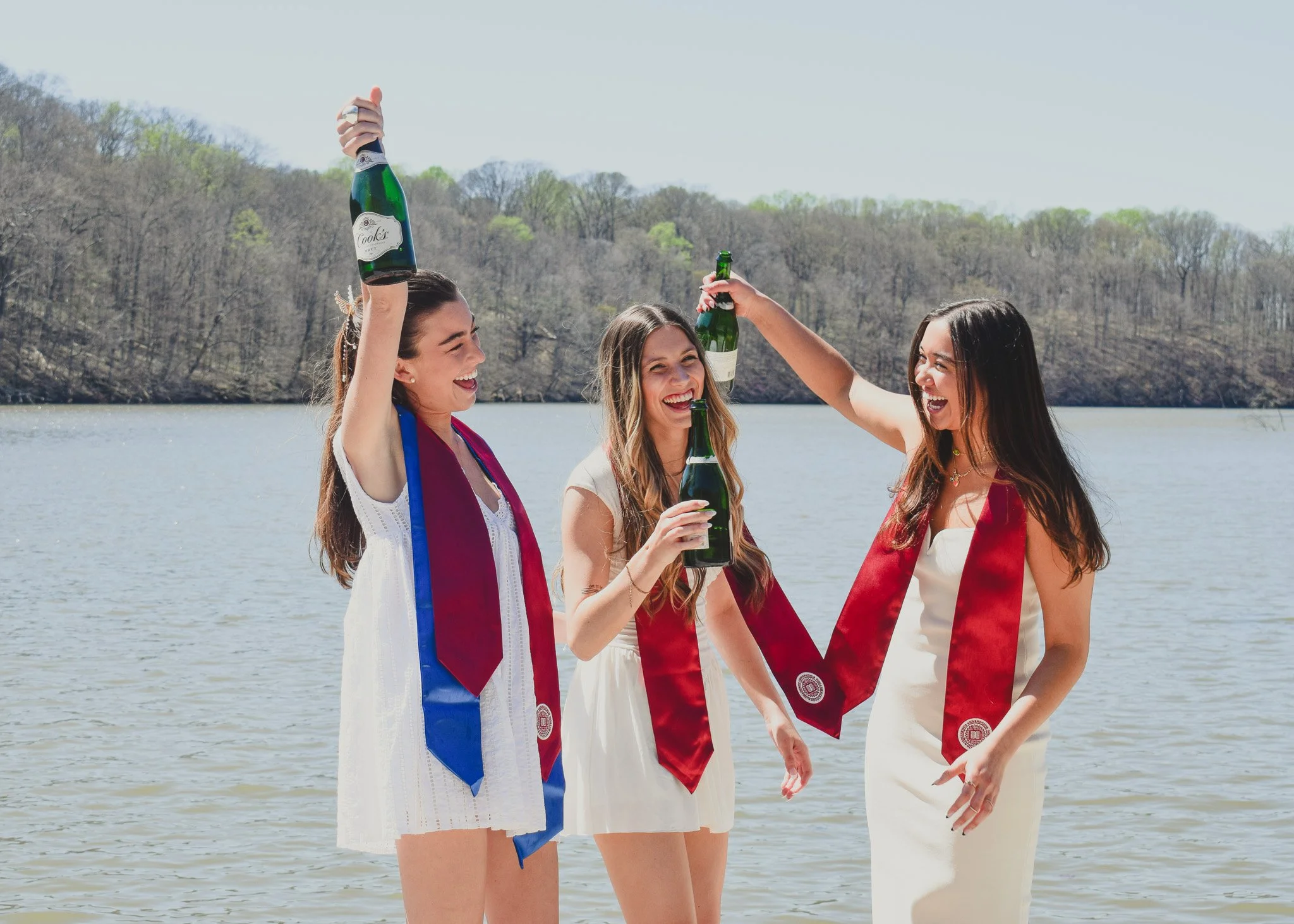 Three young women in white dresses celebrating by holding bottles of champagne and wearing maroon and blue honor cords near a lake with trees in the background.