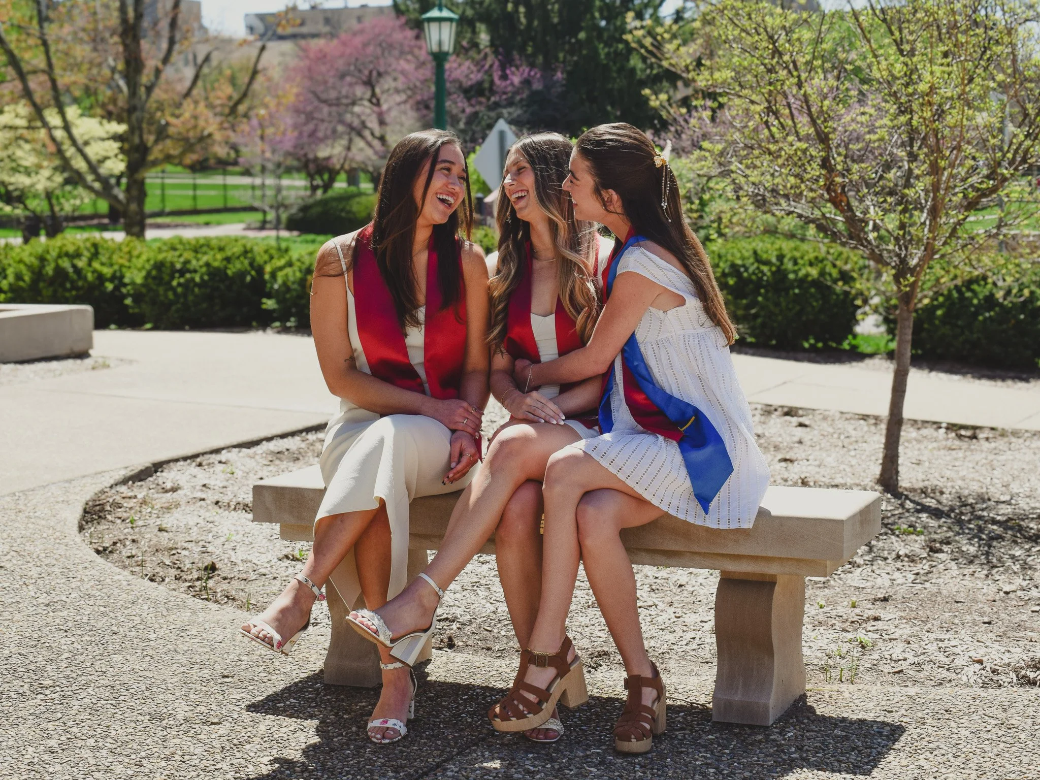 Three young women celebrating graduation sitting on a bench outdoors, dressed in graduation gowns and caps, smiling and laughing together on a sunny day.