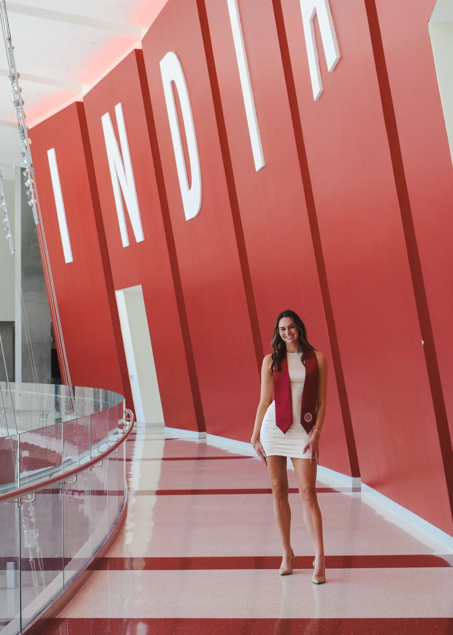 A woman standing in front of a large red wall with the partial word 'INDI' in white letters, inside a modern building with a glass railing and polished floor.