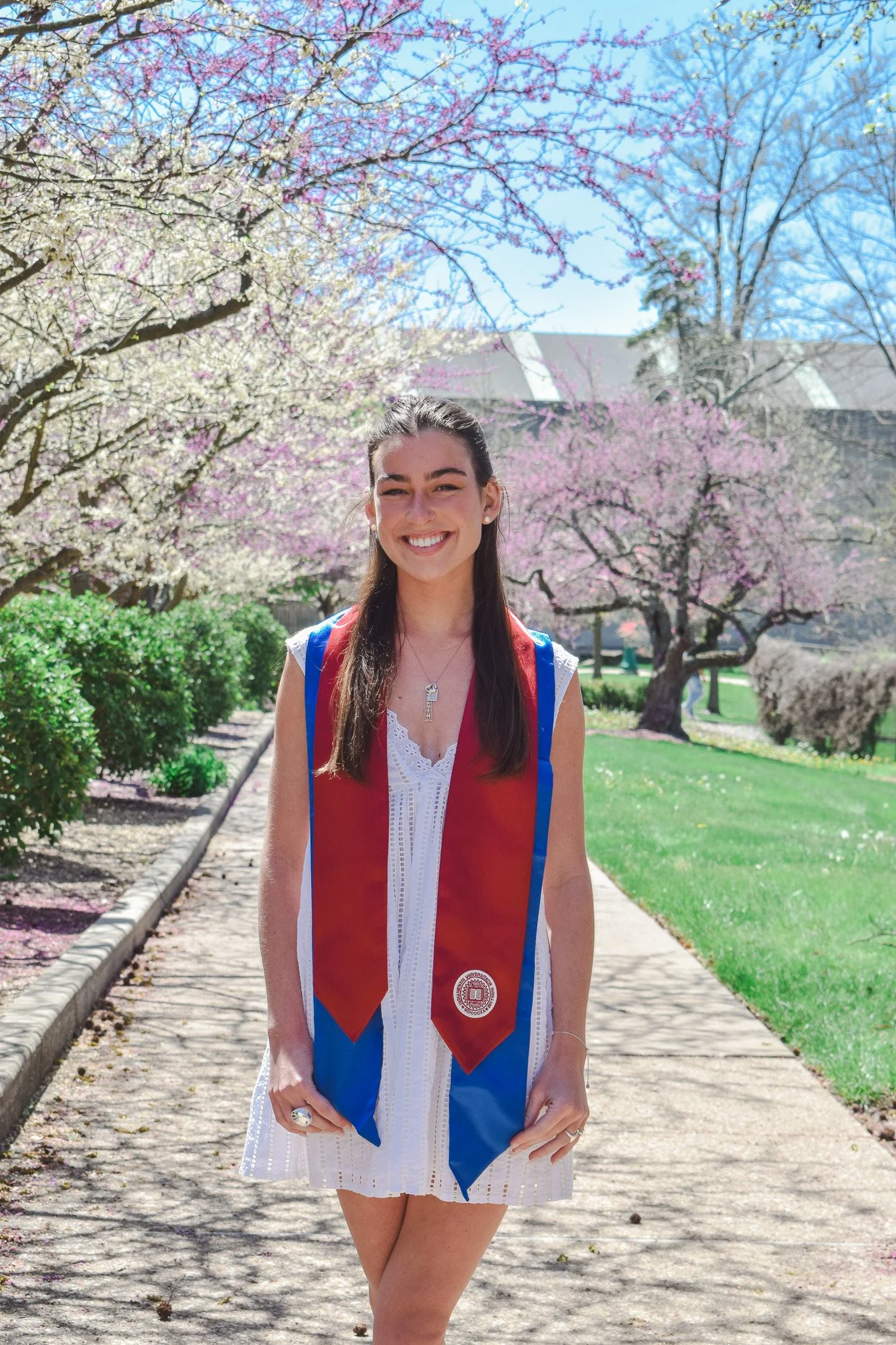 Young woman in a white dress with a red and blue graduation stole, standing on a sidewalk with blooming trees and green grass in the background.