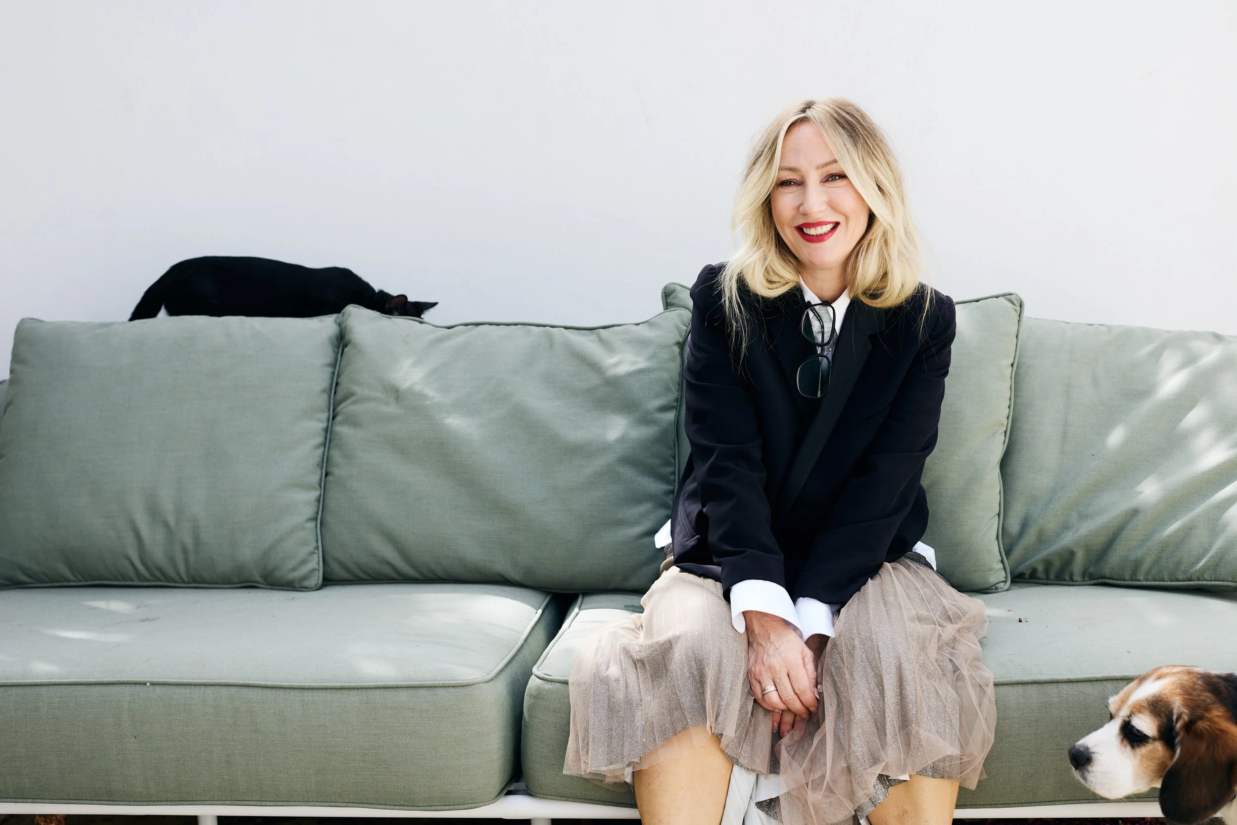 A smiling woman with blonde hair wearing a black blazer and beige skirt sitting on a light-colored sofa, with a black cat on the sofa and a dog in front of her, against a plain white background.