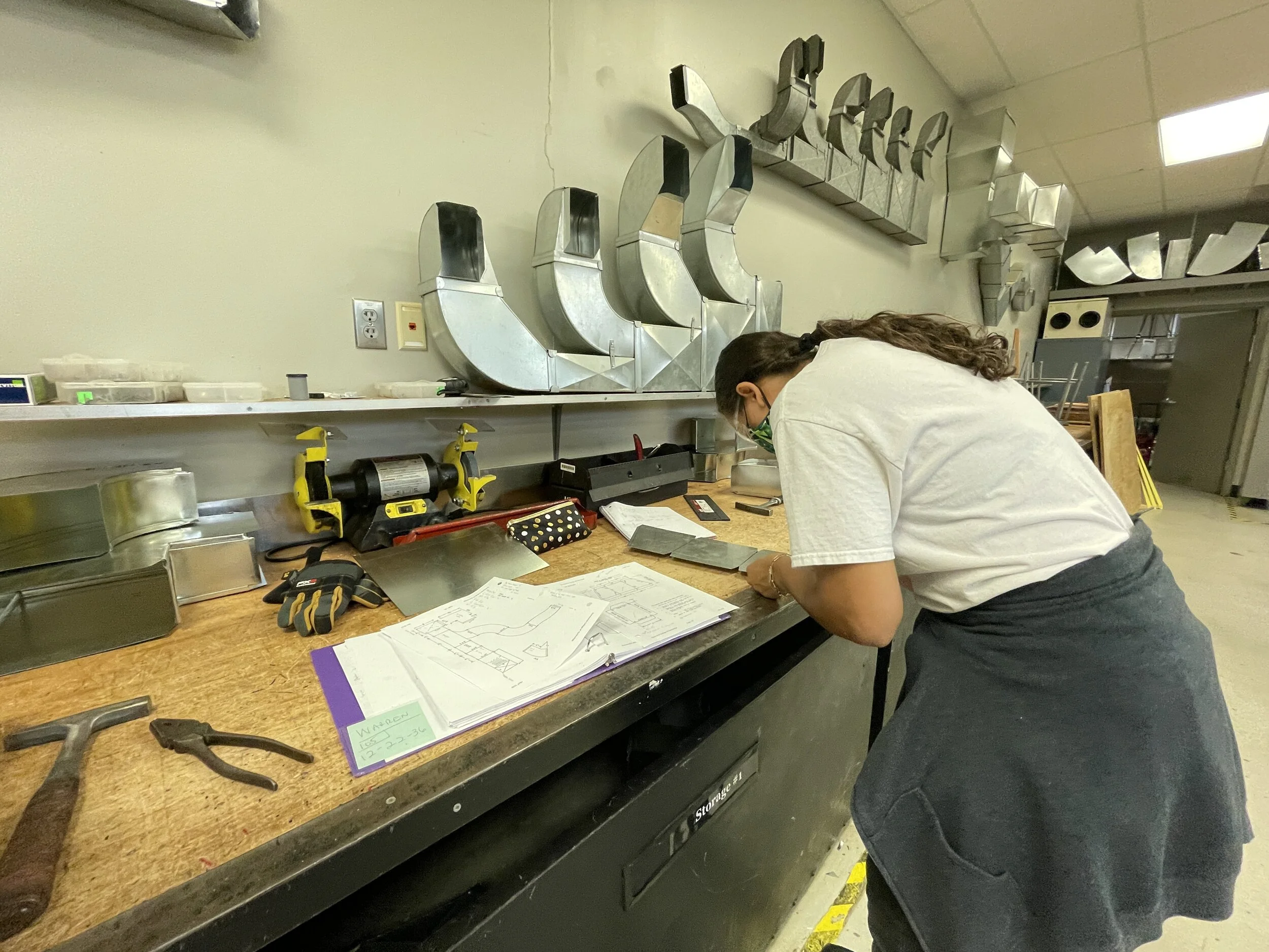 A010 - Overhead boom down on HVAC student 
ACTION: Student checking plans to her left, then making the precise cut on the sheet metal