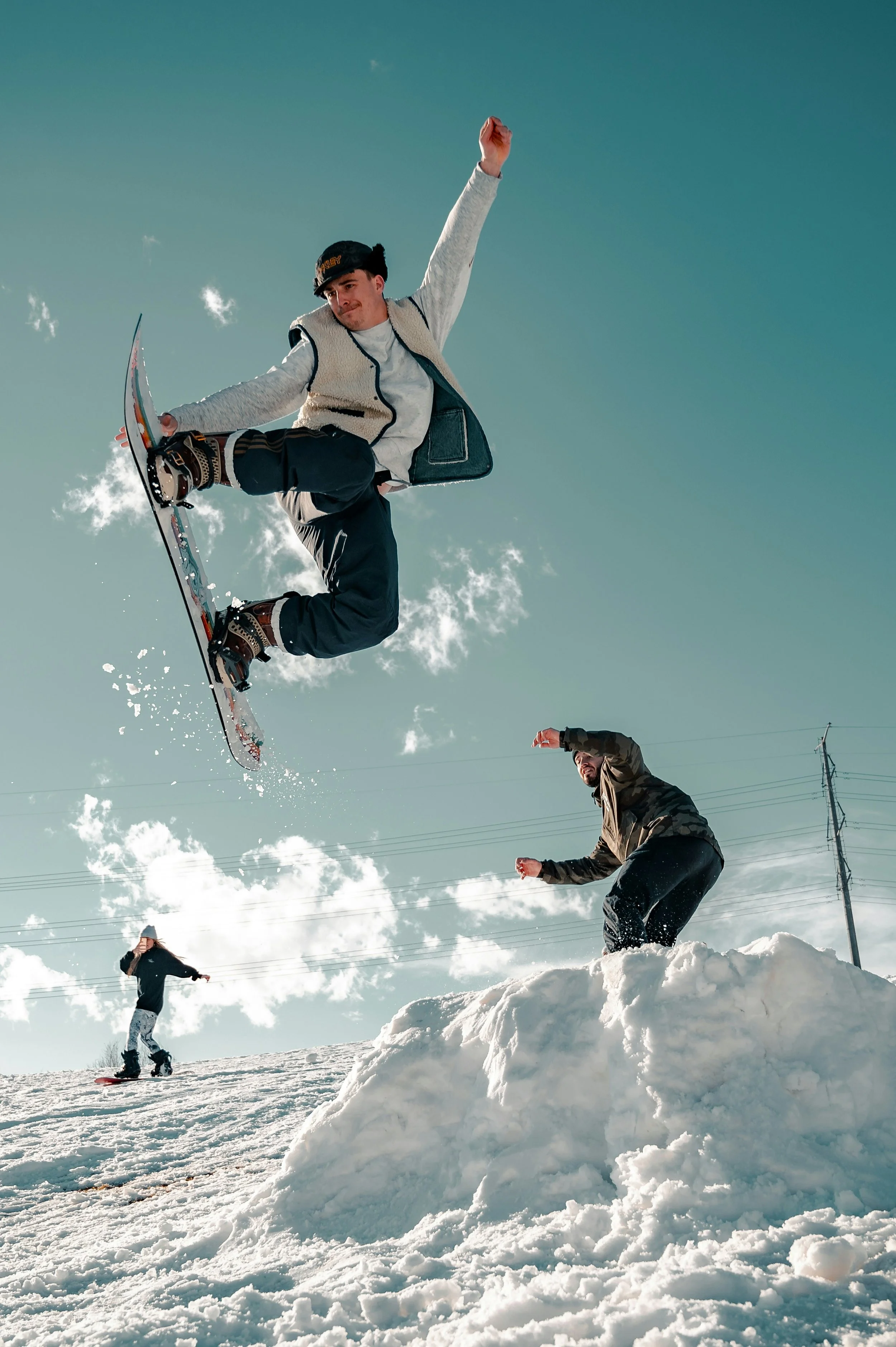 3 snowboarders with one in the air doing a trick jump and another male snowboarder about to do a jump over a mound of snow