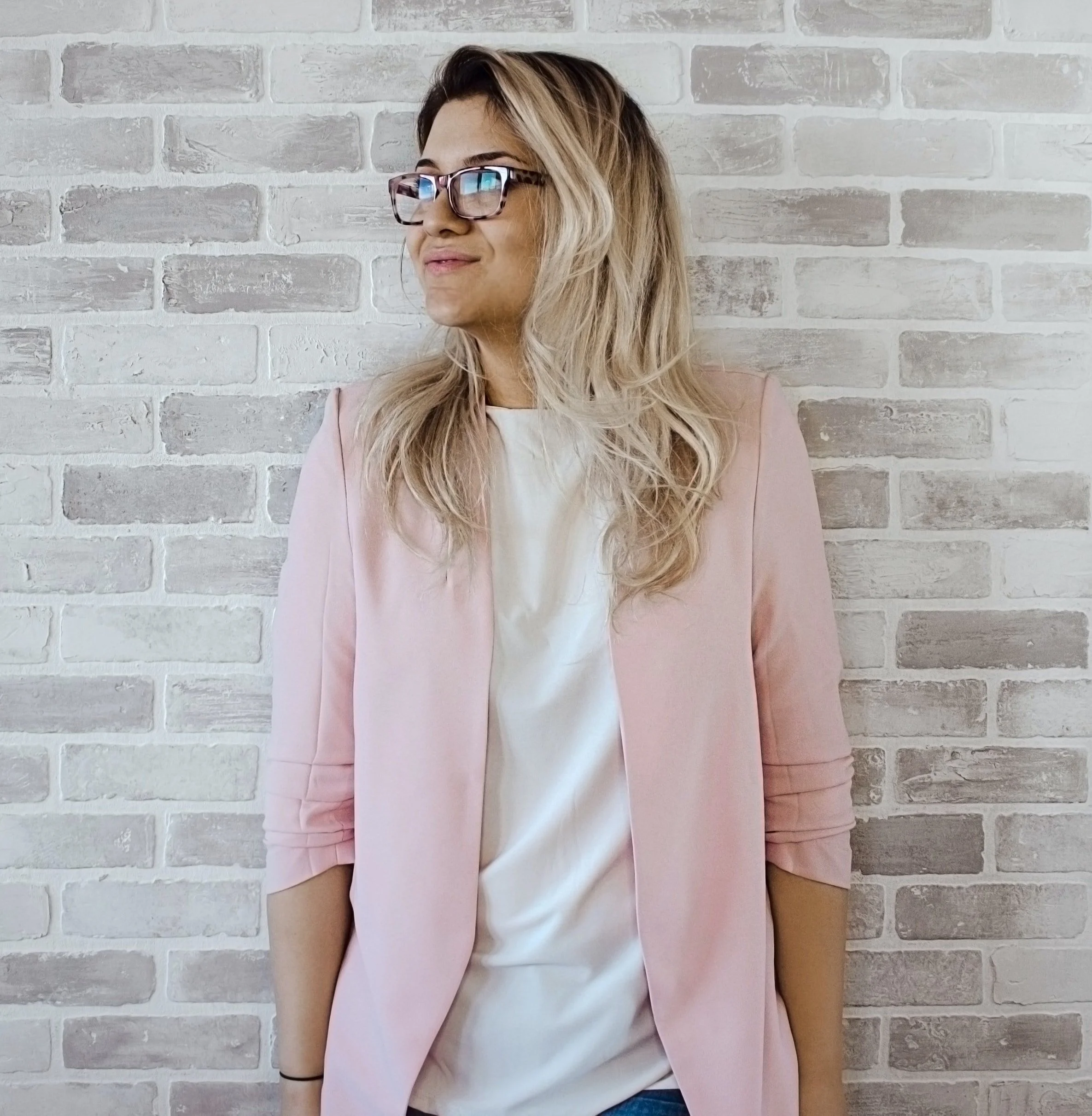 A woman with long blonde hair, wearing glasses, a light pink blazer, and a white top, standing against a white and gray brick wall.