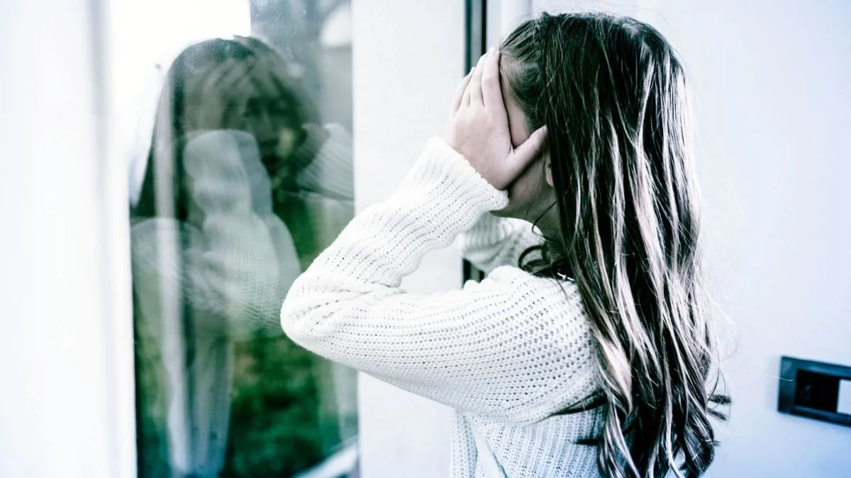 A girl with long hair covering her face with her hands, standing next to a glass window and looking out, appearing distressed or crying.