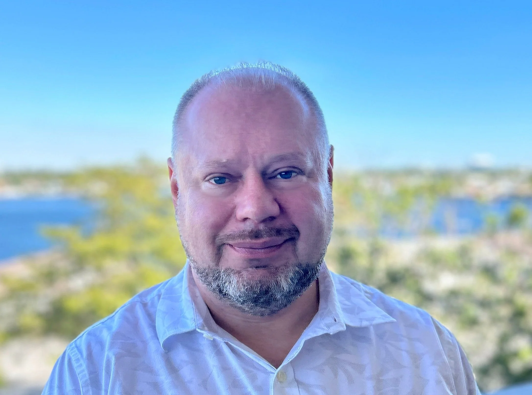 A man with a beard and short hair wearing a white shirt, standing outdoors with a cityscape and river in the background.