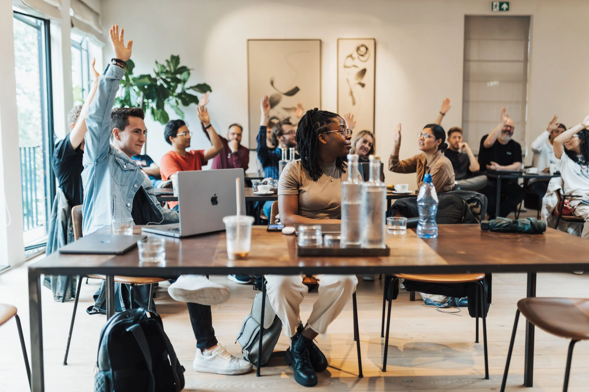 A diverse group of people in a classroom or conference room, some raising their hands, engaged in a discussion or presentation, with laptops, notebooks, and beverages on the table.
