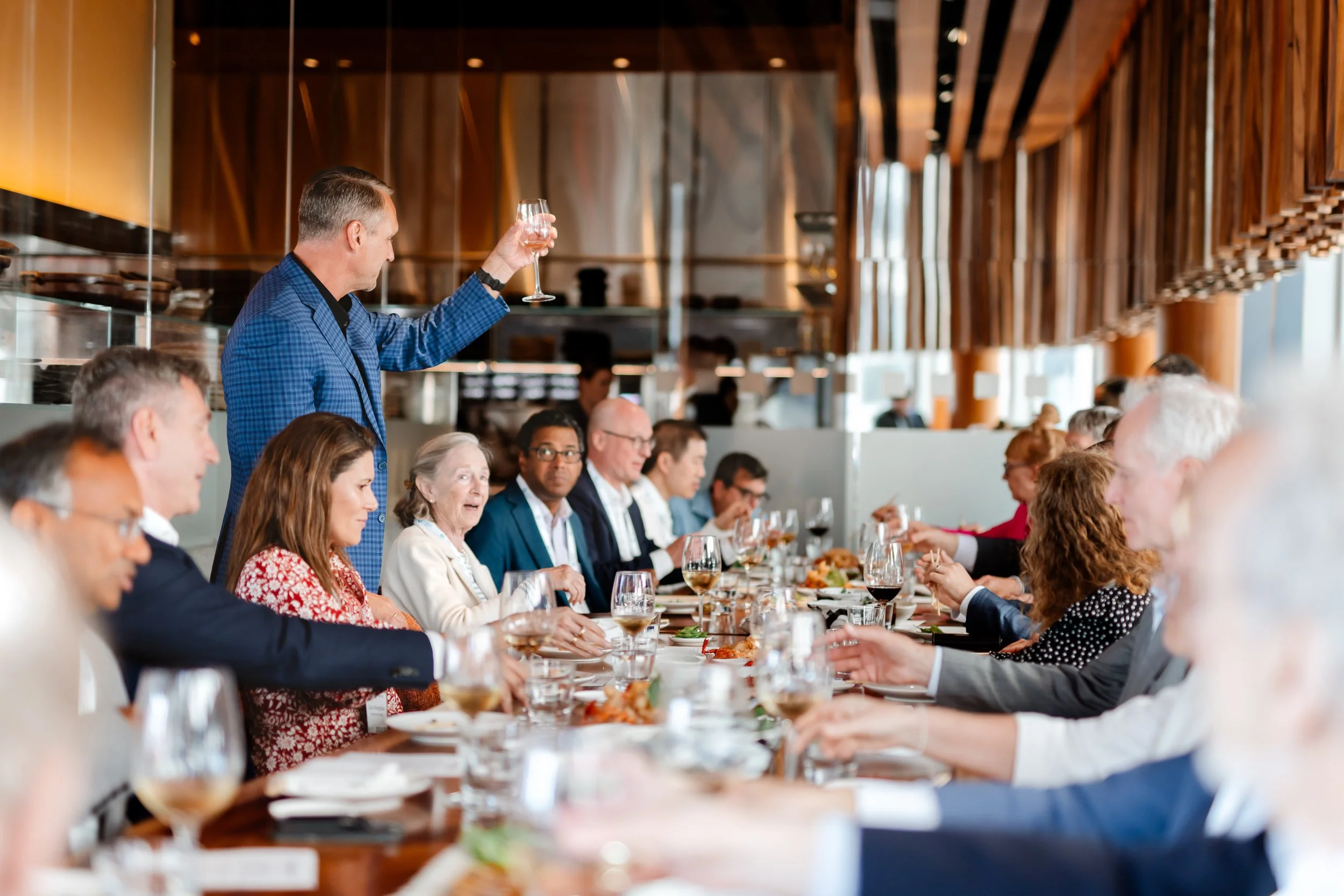 A man in a blue checkered suit raising a glass at a formal dining event while others are seated at a long table in a modern restaurant with wooden decor.