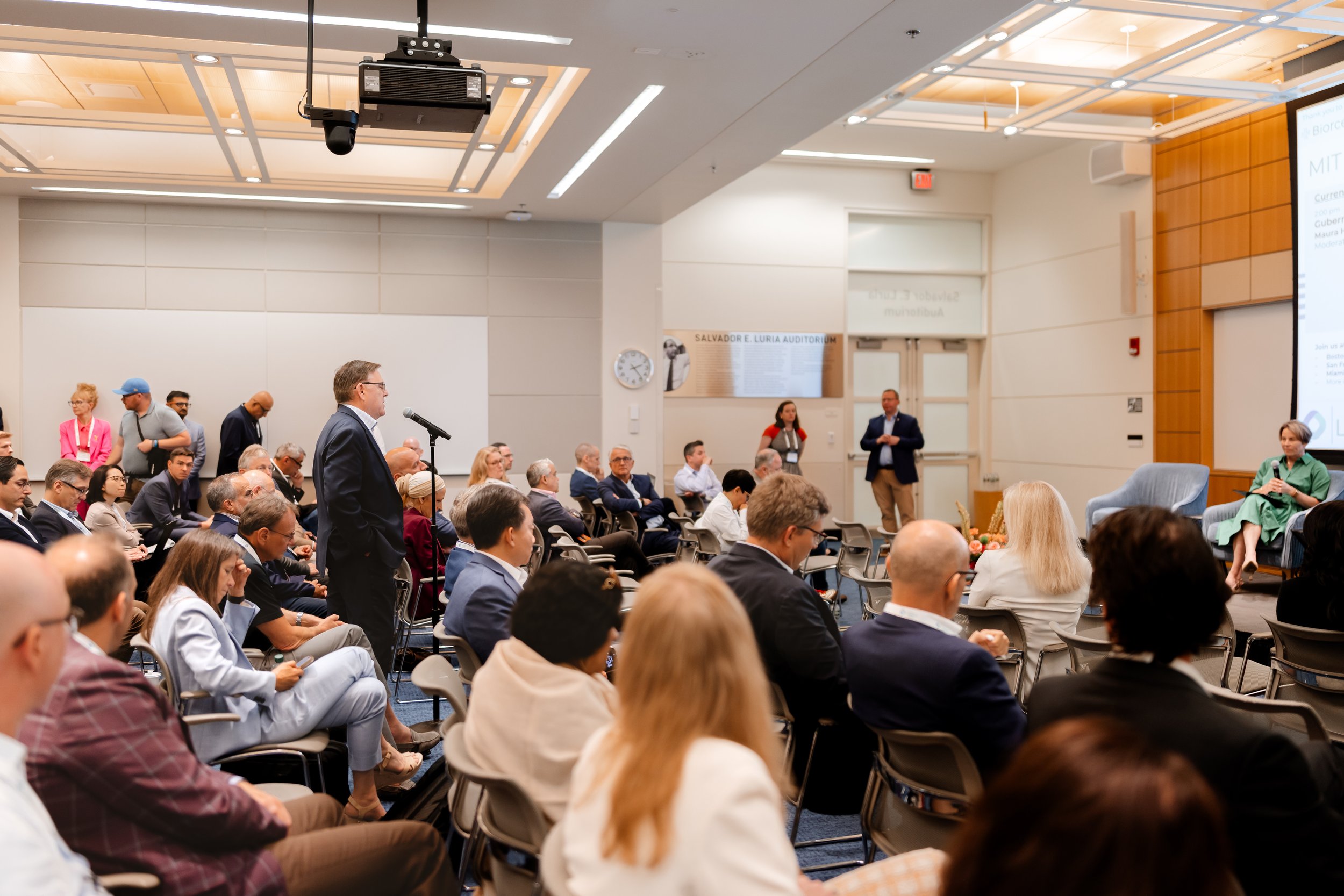 A conference room filled with seated people listening to a man at a microphone. A large presentation screen is visible on the right side of the image. Some attendees are taking notes, and others are listening attentively. The room has a high ceiling with modern lighting.