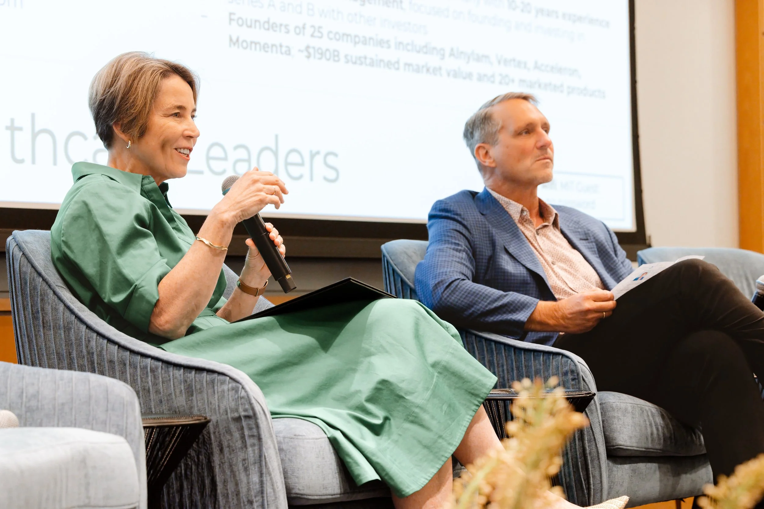 A woman in a green dress speaking into a microphone during a panel discussion, seated next to a man in a blue plaid suit reading a pamphlet, with a presentation screen in the background. Governor Maura Healey of Massachusetts