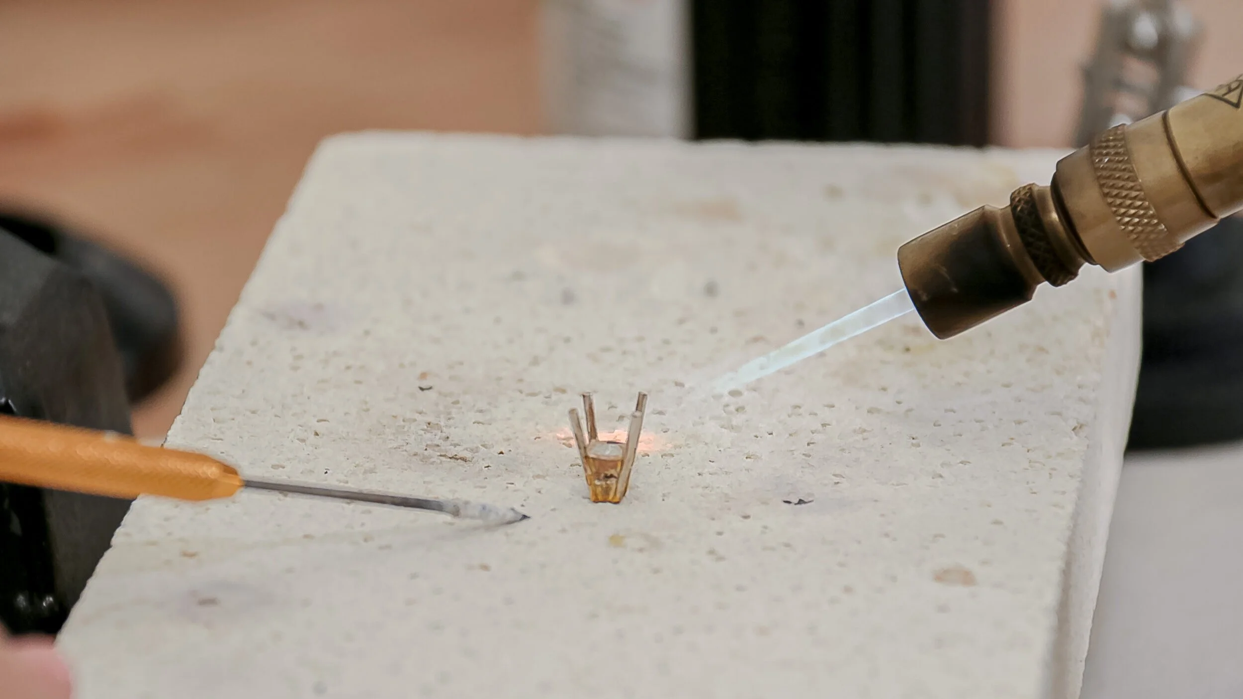 Jewellery making process soldering onto a small metal structure on a ceramic tile surface, using a handheld torch with a flame.