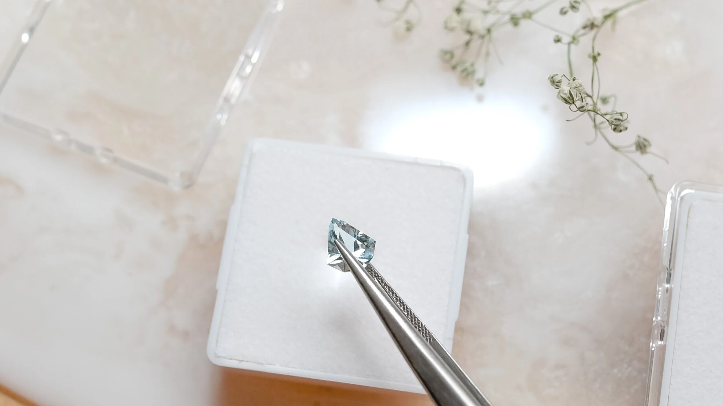 A close-up of a clear, faceted gemstone held with metal tweezers, placed on a white cushion in a display box, with glass display cases and a sprig of small white flowers visible in the background.